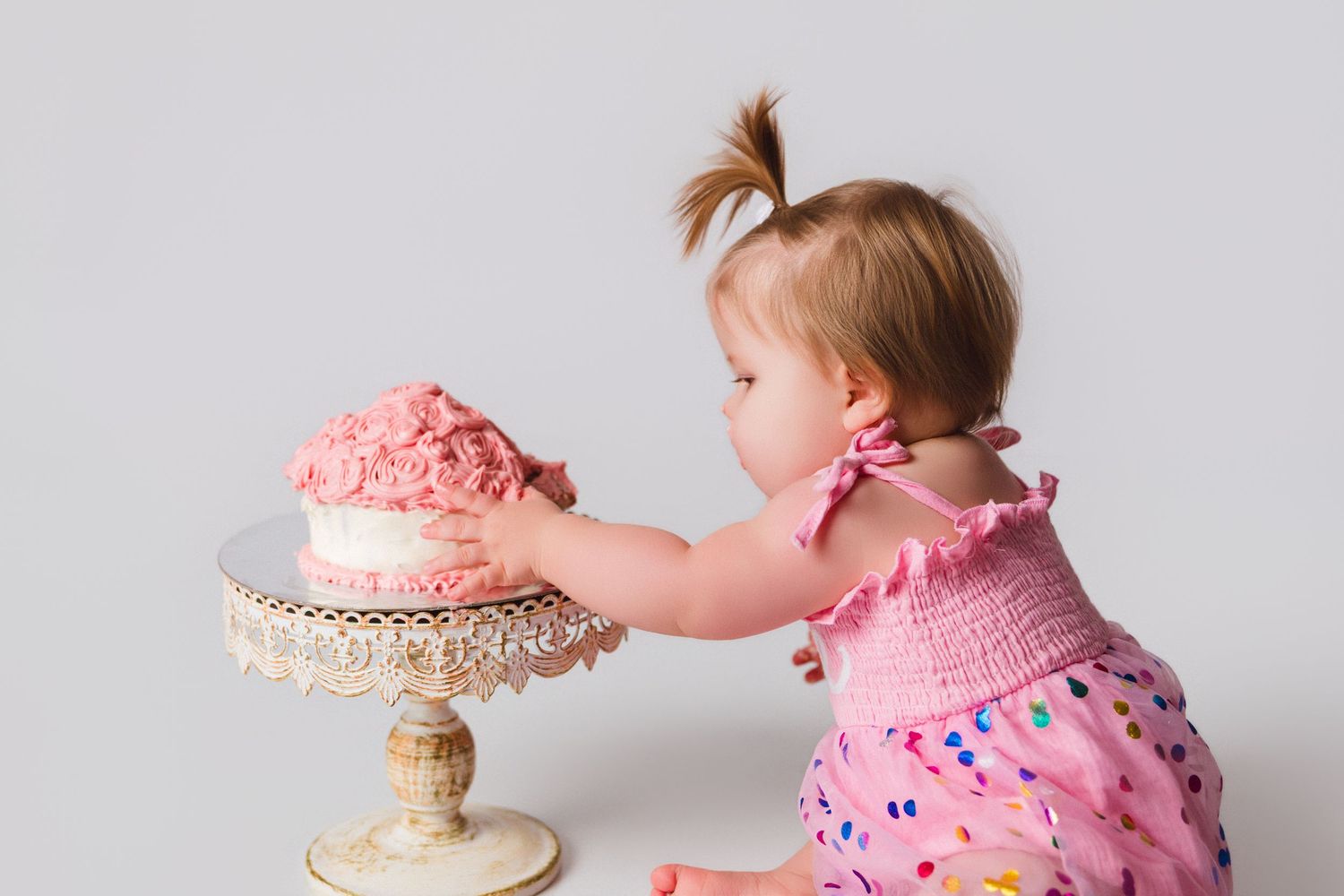 Baby in pink polka dot dress explores cake during first birthday photoshoot.