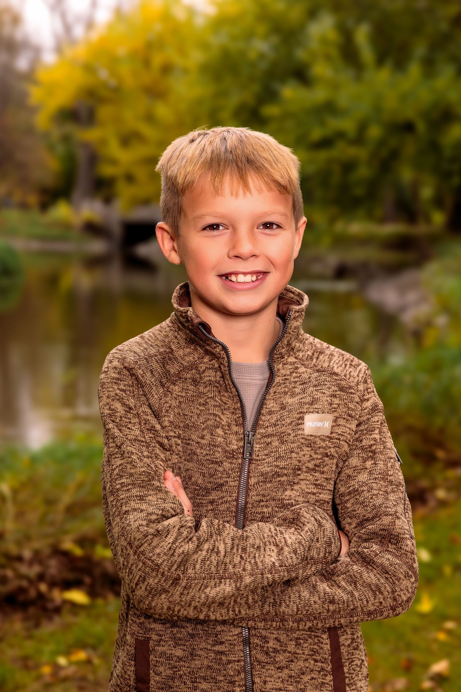Young child wearing brown sweater smiling outdoors in autumn setting with fall foliage in background.