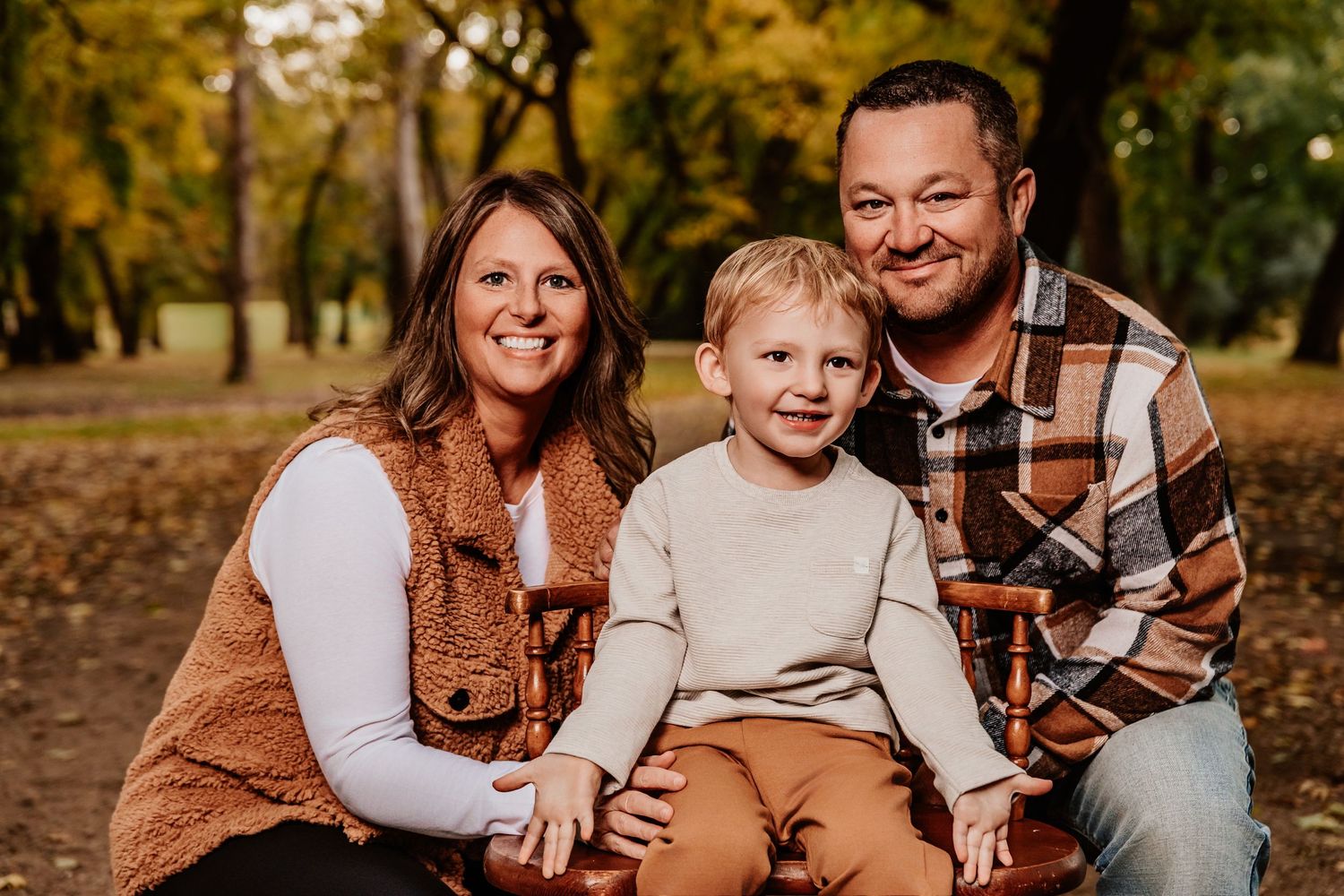 A family of three in casual fall attire poses together on a wooded trail with autumn foliage.