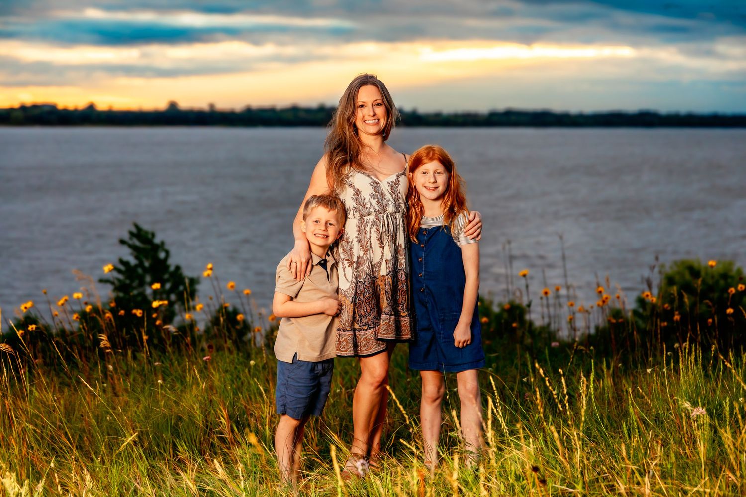 Three people stand together against a dramatic sunset sky over a waterfront.