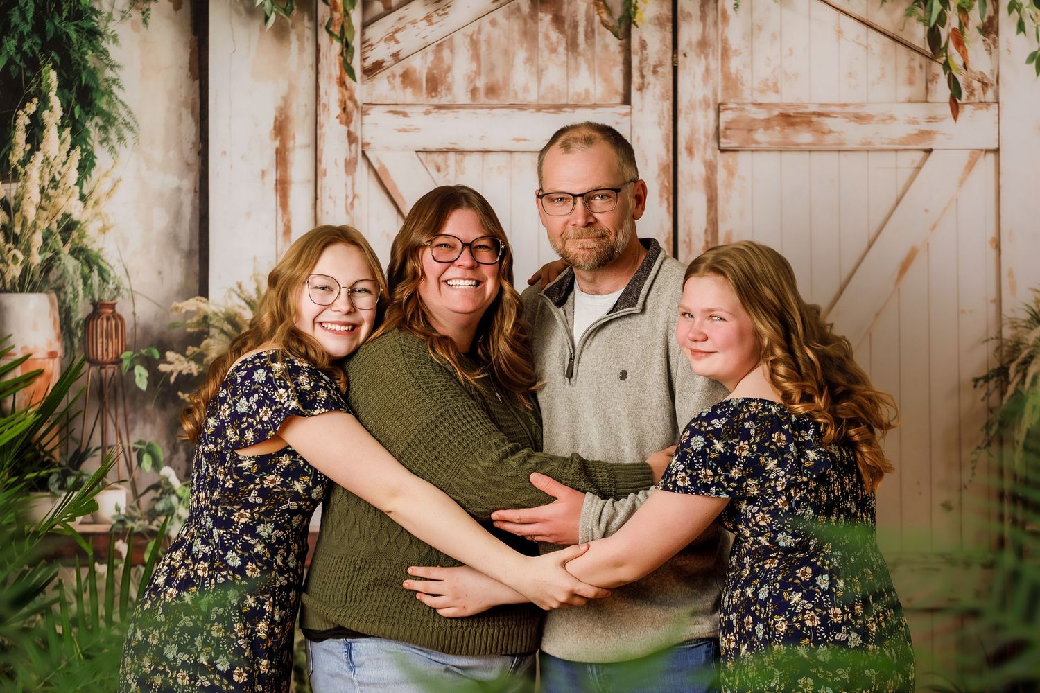 Family of four smiling and embracing in front of rustic barn doors with lush greenery backdrop.