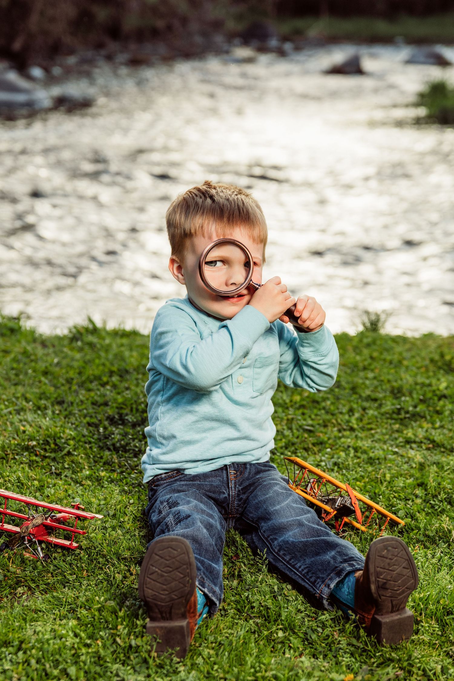 Young boy sitting on grass by a river, looking through a magnifying glass with toy planes beside him.