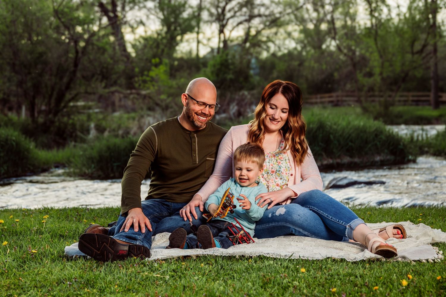 Happy family with baby sitting on blanket by river in green park during golden hour outdoor photo session.