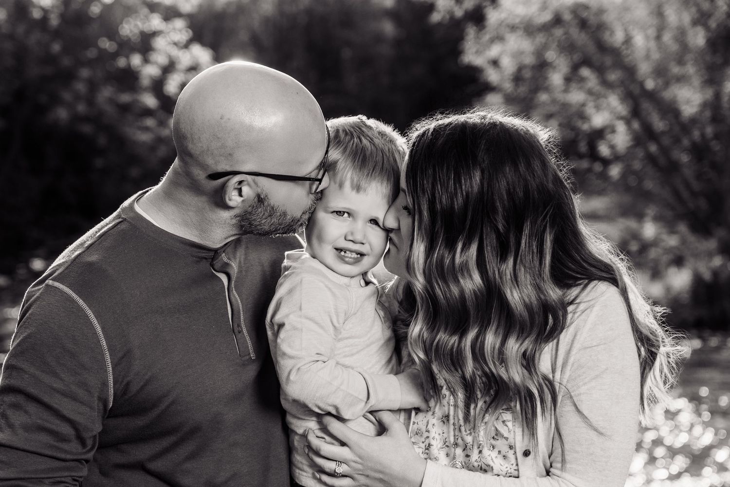 Black and white photo of a bald father and brunette mother kissing their smiling toddler outdoors.