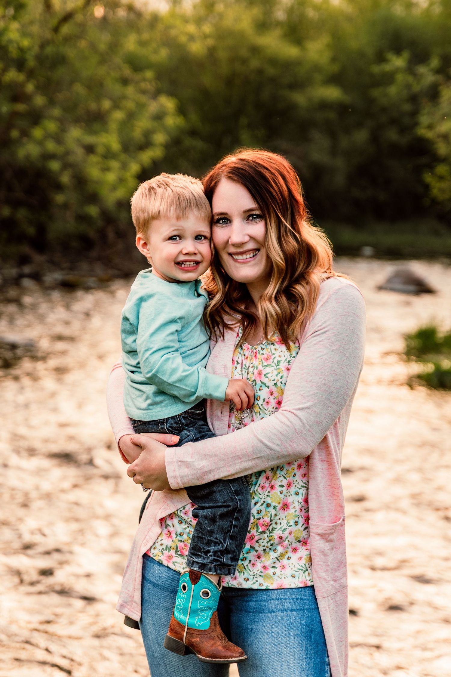 A smiling redhead woman holds a happy toddler boy in cowboy boots outdoors near a sandy riverbank.