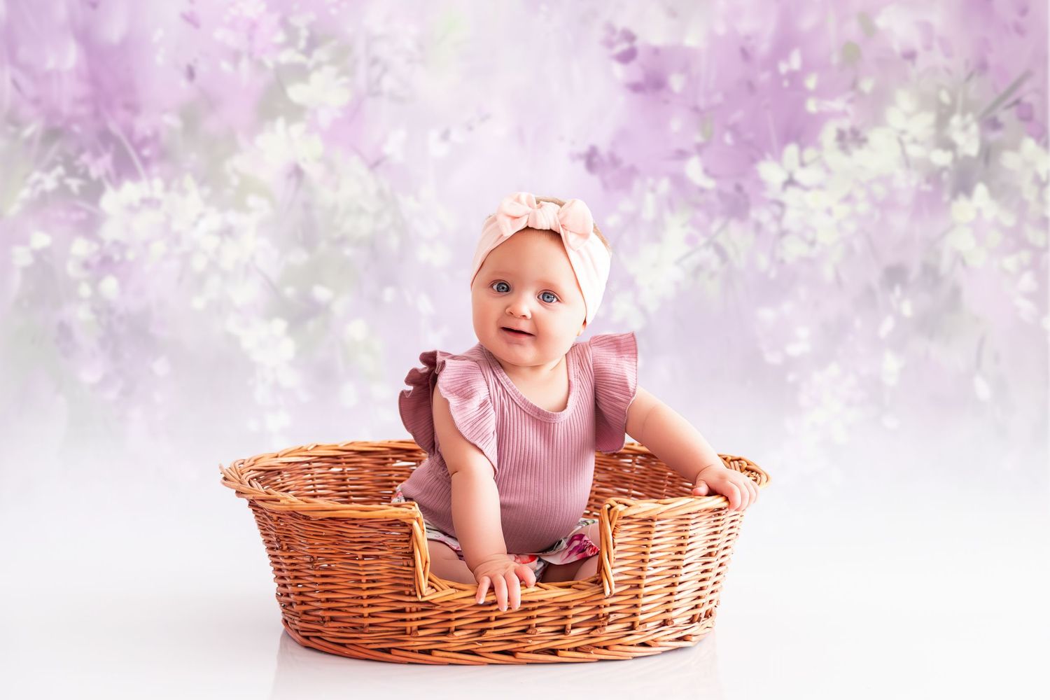 A baby in a pink dress sits in a woven wicker basket surrounded by soft pink floral effects.