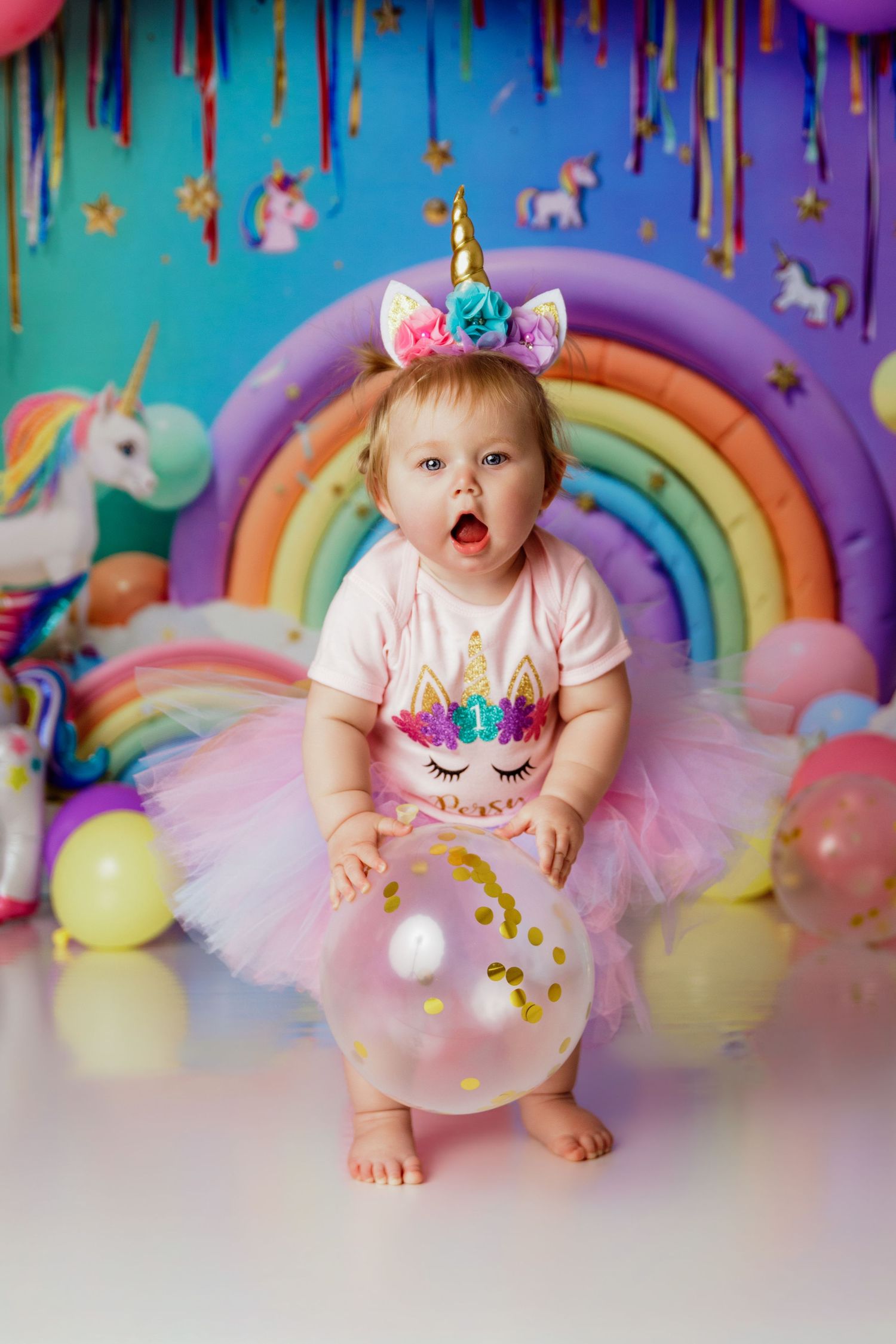 Cute baby girl in unicorn tutu outfit and headband at colorful rainbow first birthday party.