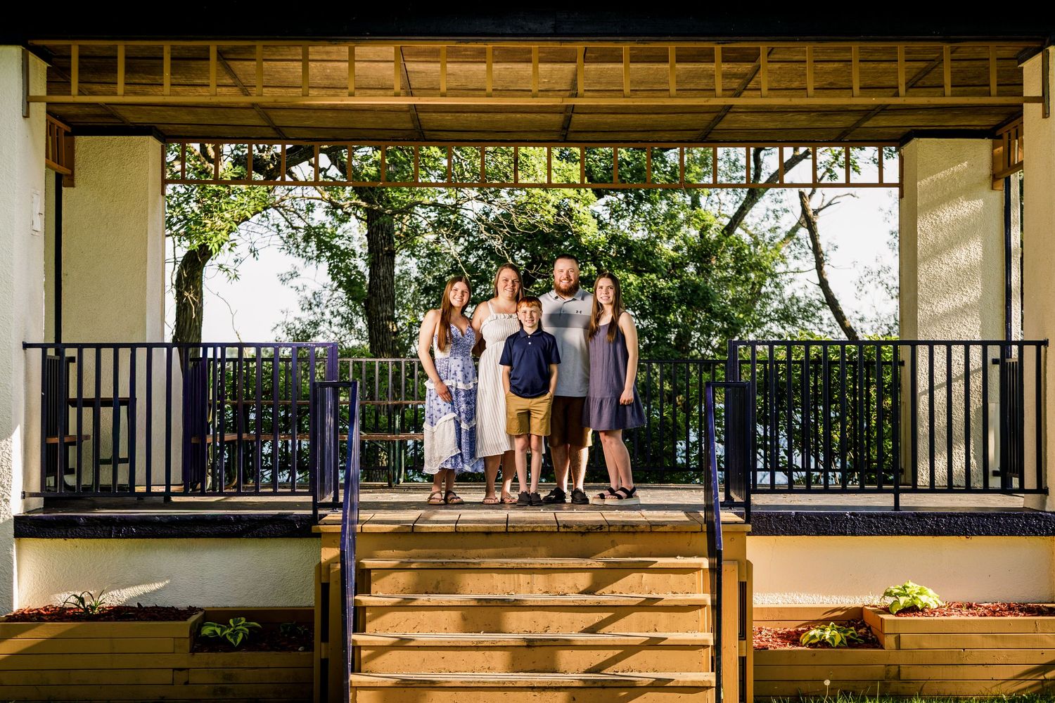 Family of five standing together on a wooden deck surrounded by lush green trees near a lake.