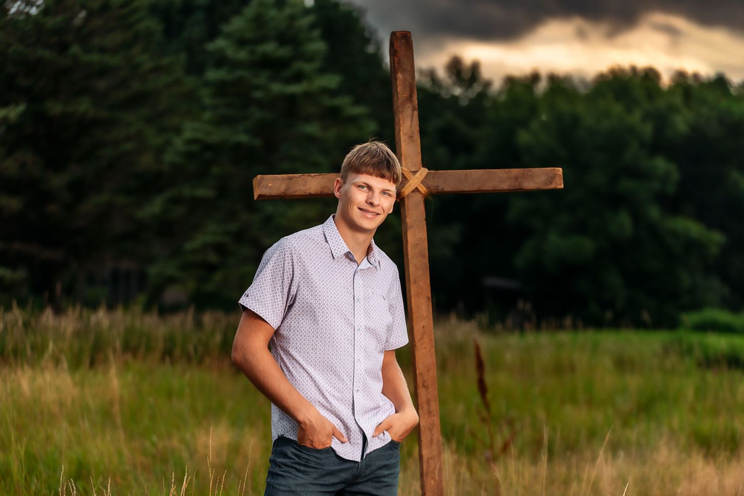 Young man leaning against a rustic wooden cross in a field with dramatic cloudy sky backdrop.