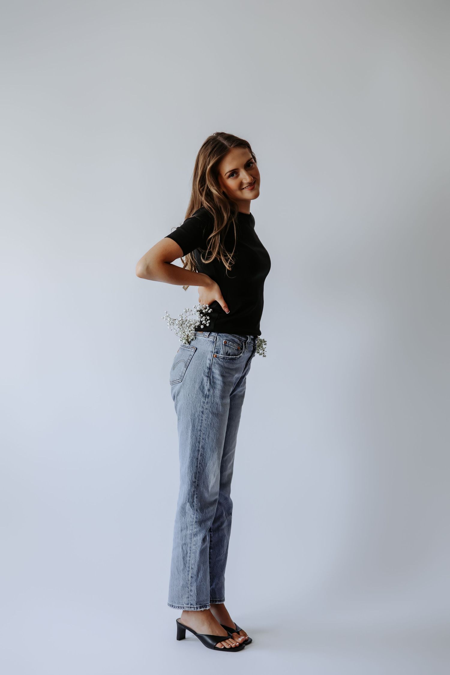 Woman in black tee and jeans posing sideways holding small white flowers, studio shot.