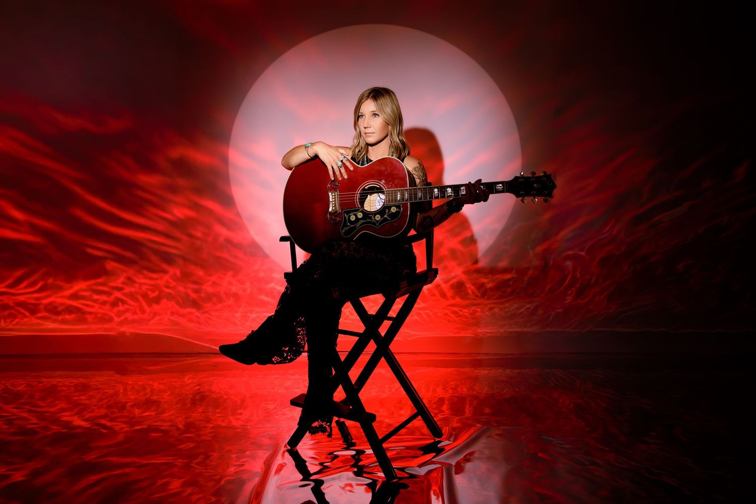 Blonde woman sitting on a director's chair holding an acoustic guitar against a dramatic red-lit background.