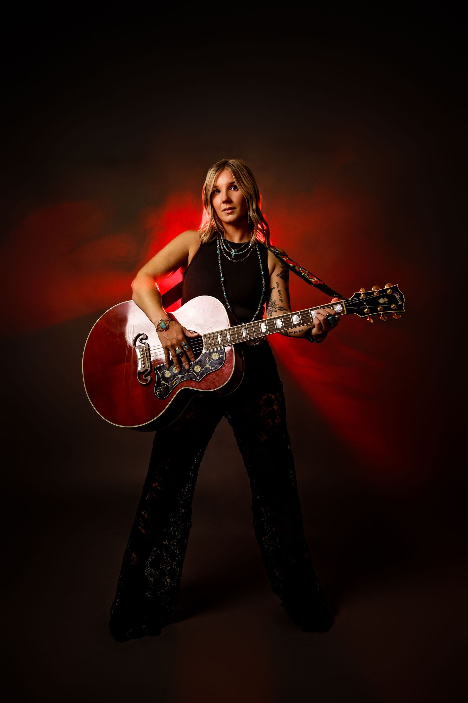 Tattooed female musician standing with red Gibson acoustic guitar against dark red dramatic backdrop.