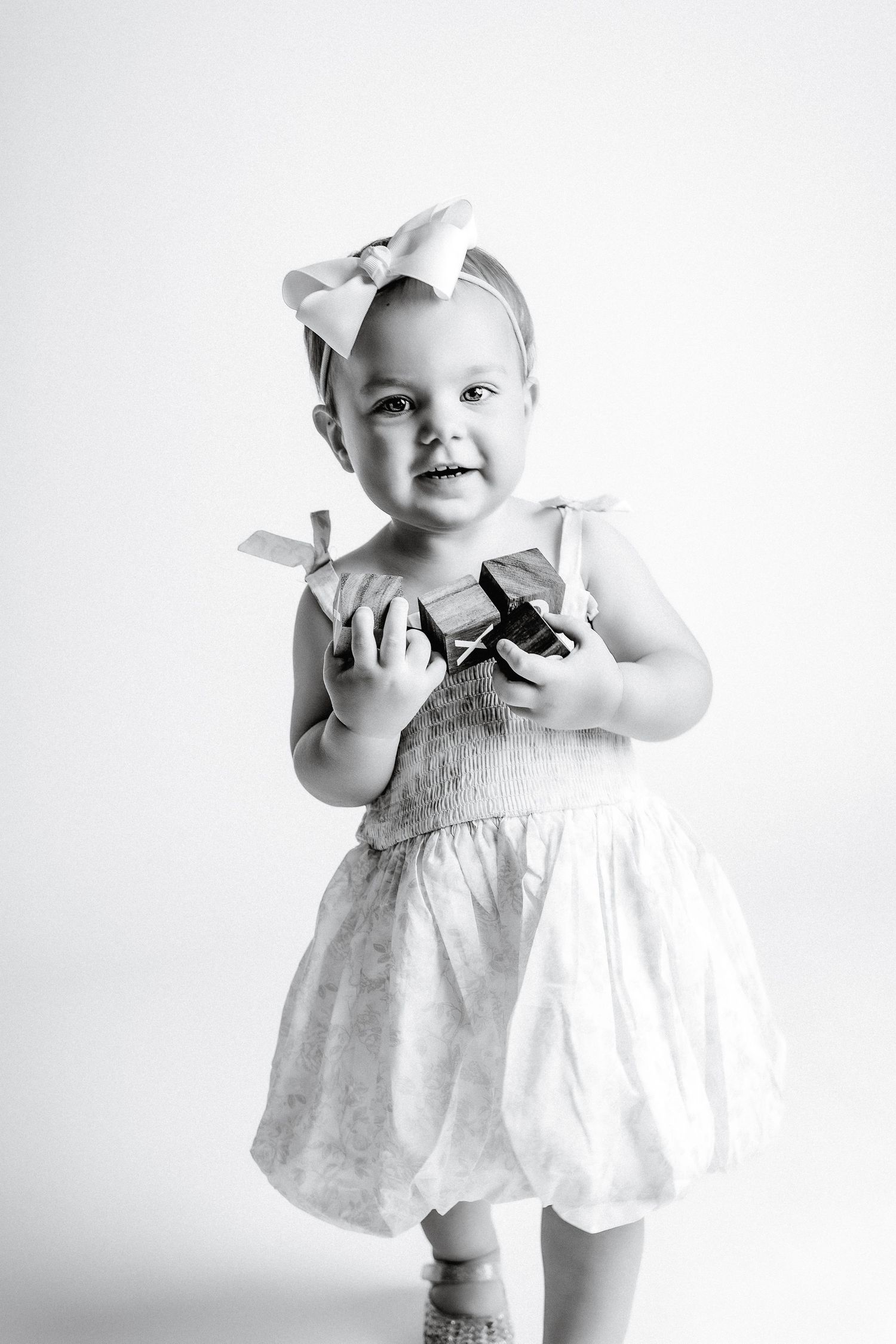 Black and white photo of a smiling toddler girl in a dress and bow headband holding small gift boxes.