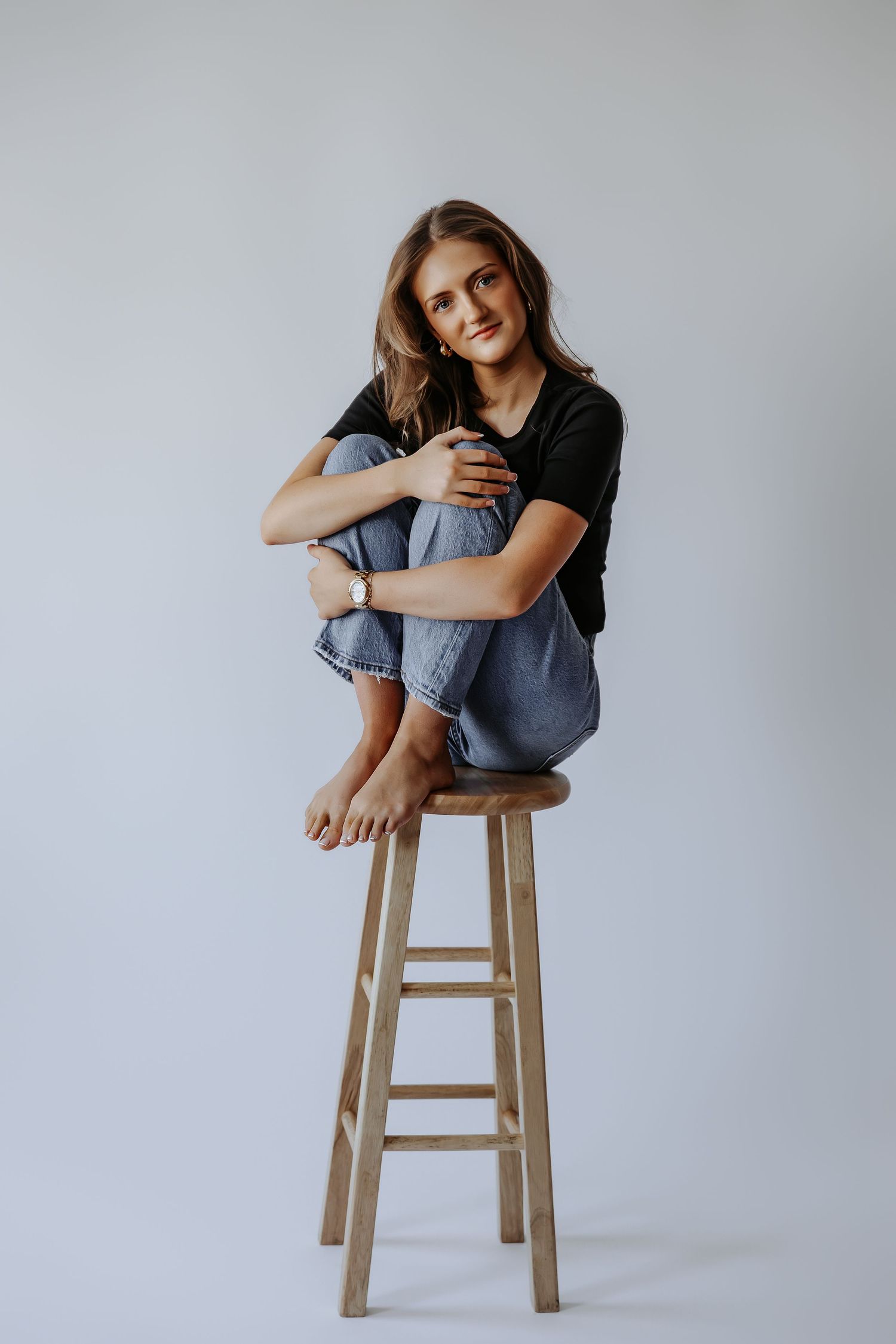 Young woman in black tee and jeans sits cross-legged on wooden stool, smiling at camera against gray background.