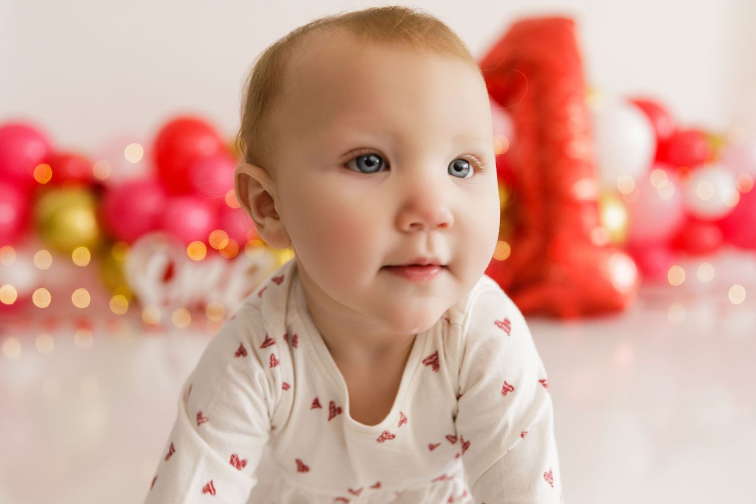 Close-up portrait against red and gold Valentine's Day decorations with soft bokeh lights in background.