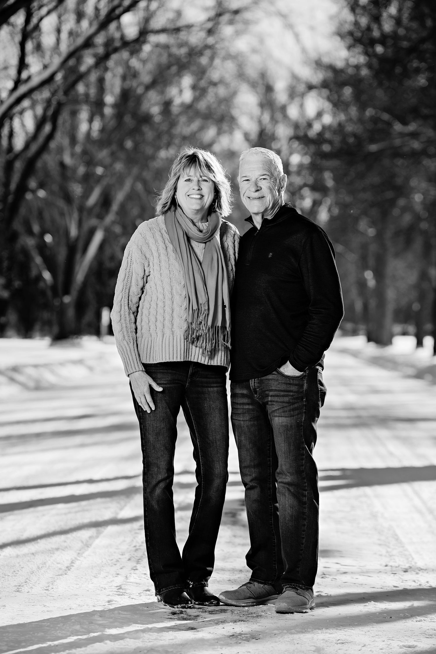 A couple stands together on a snowy path in winter wearing casual sweaters and jeans in black and white.