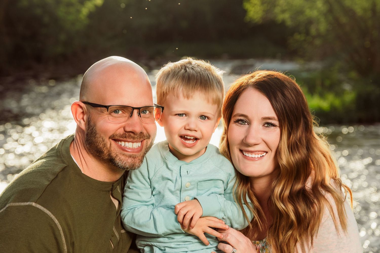 Happy family portrait of a bald father, toddler son, and brunette mother smiling outdoors near a sunlit river.
