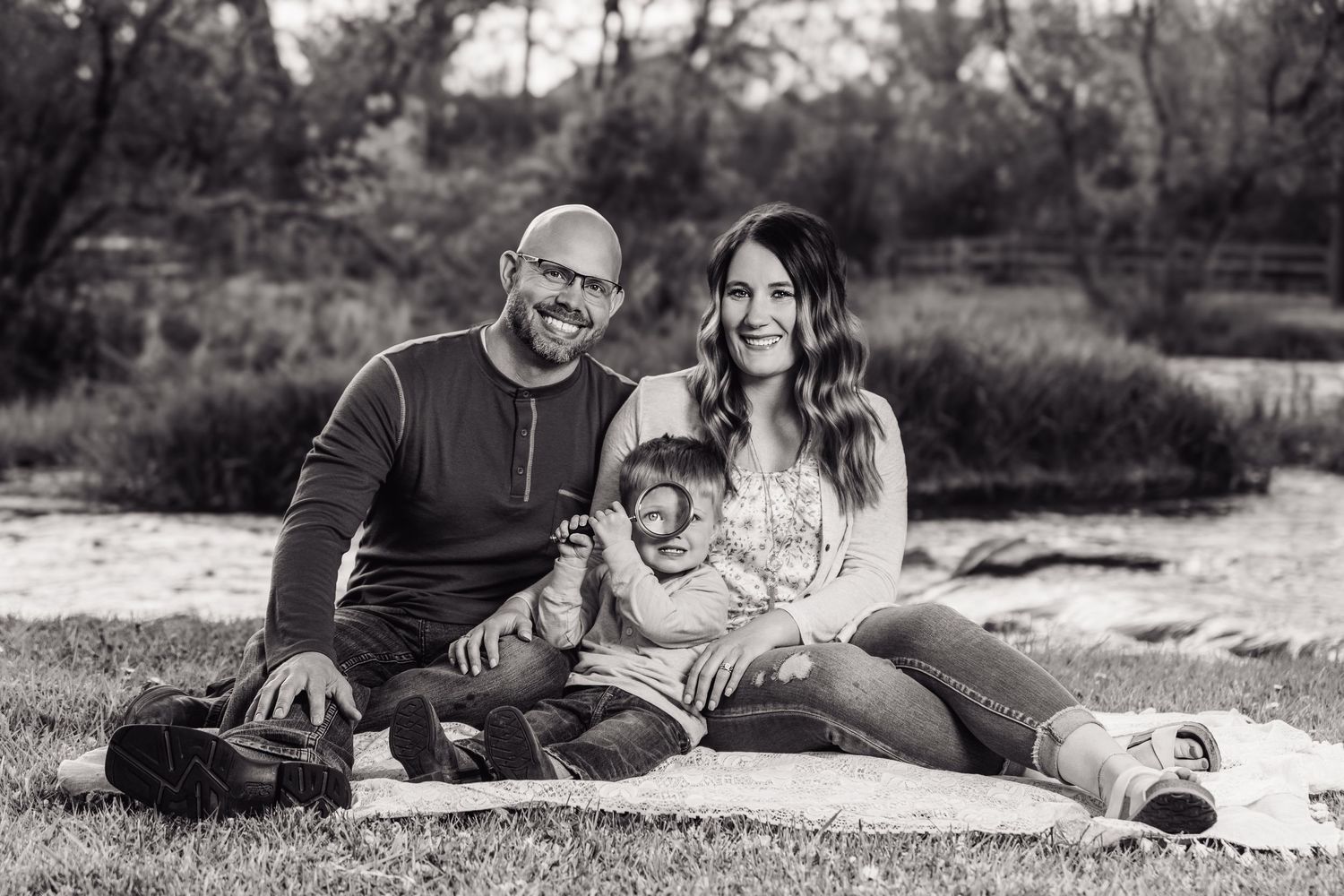 Black and white family portrait of smiling couple with baby sitting on blanket outdoors in natural setting.
