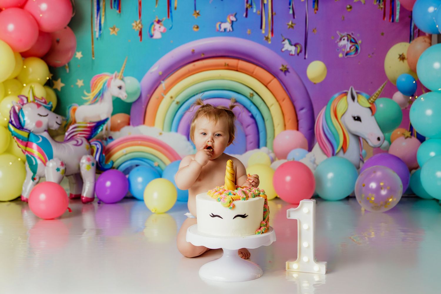 Baby girl celebrates first birthday smashing a unicorn cake surrounded by colorful balloons and rainbow decorations.