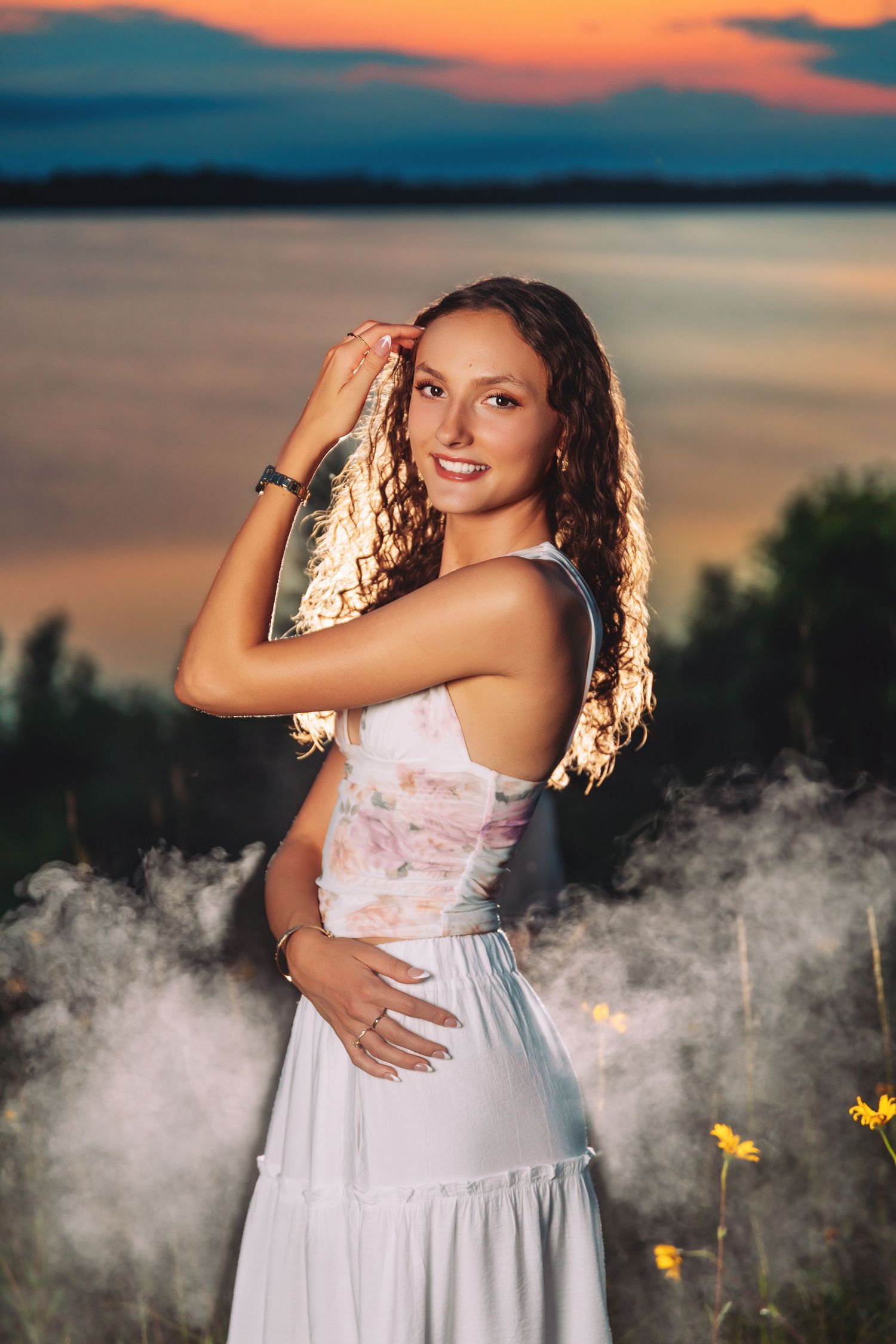 A figure in a flowing white dress poses against a dramatic sunset sky with ethereal smoke and yellow butterflies at dusk.