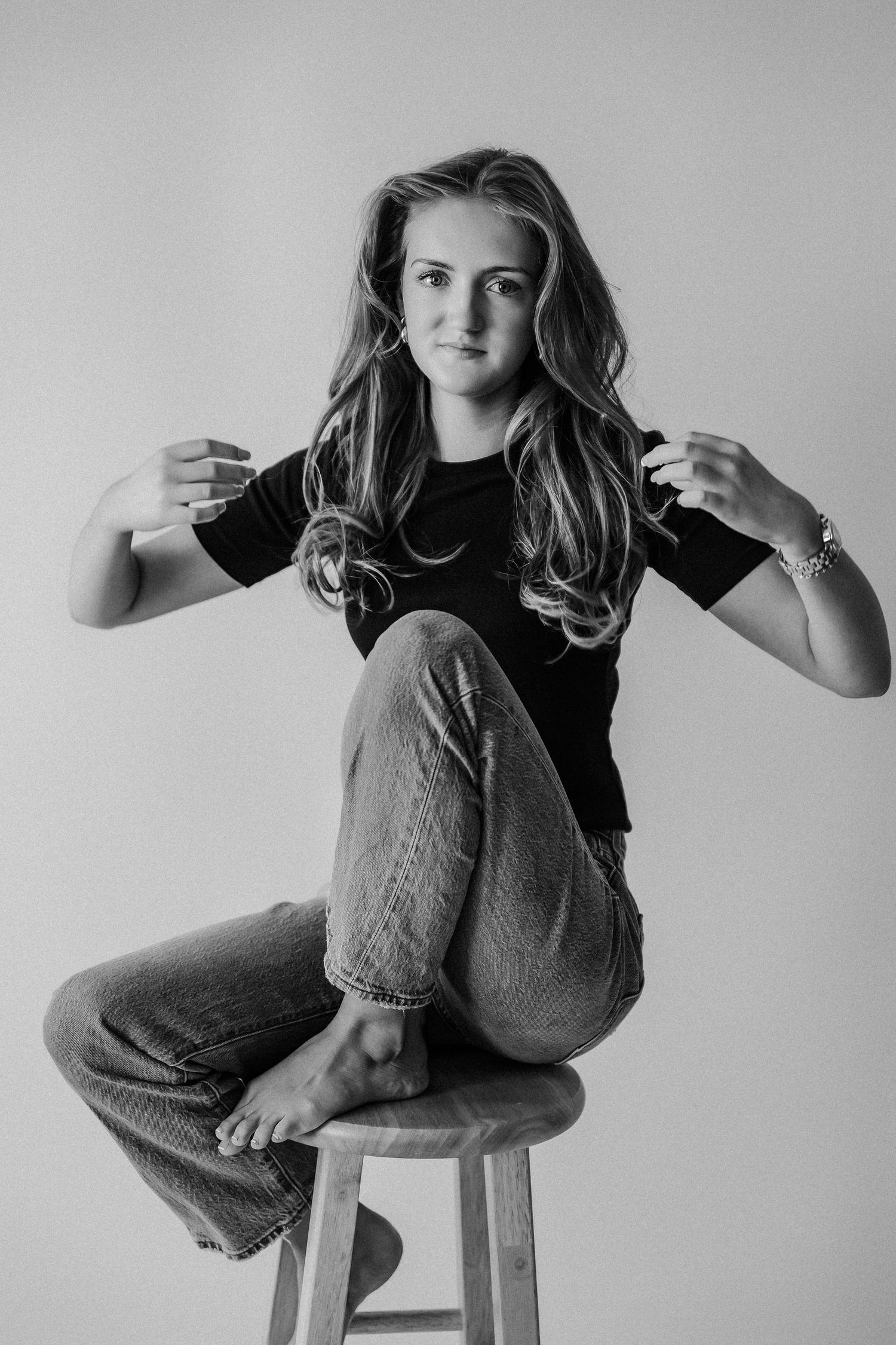 Young woman in black tee and jeans sits cross-legged on wooden stool, posing playfully in black and white studio photo.