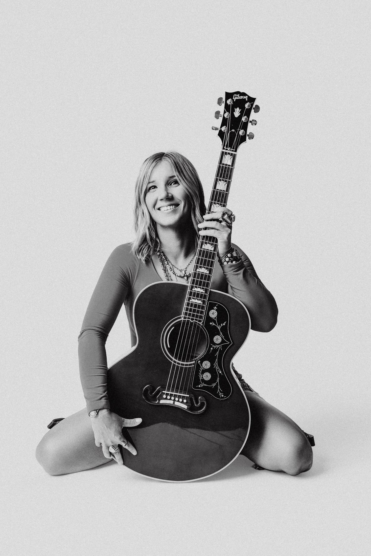 Smiling woman sitting on floor holding acoustic Gibson guitar in black and white photo.