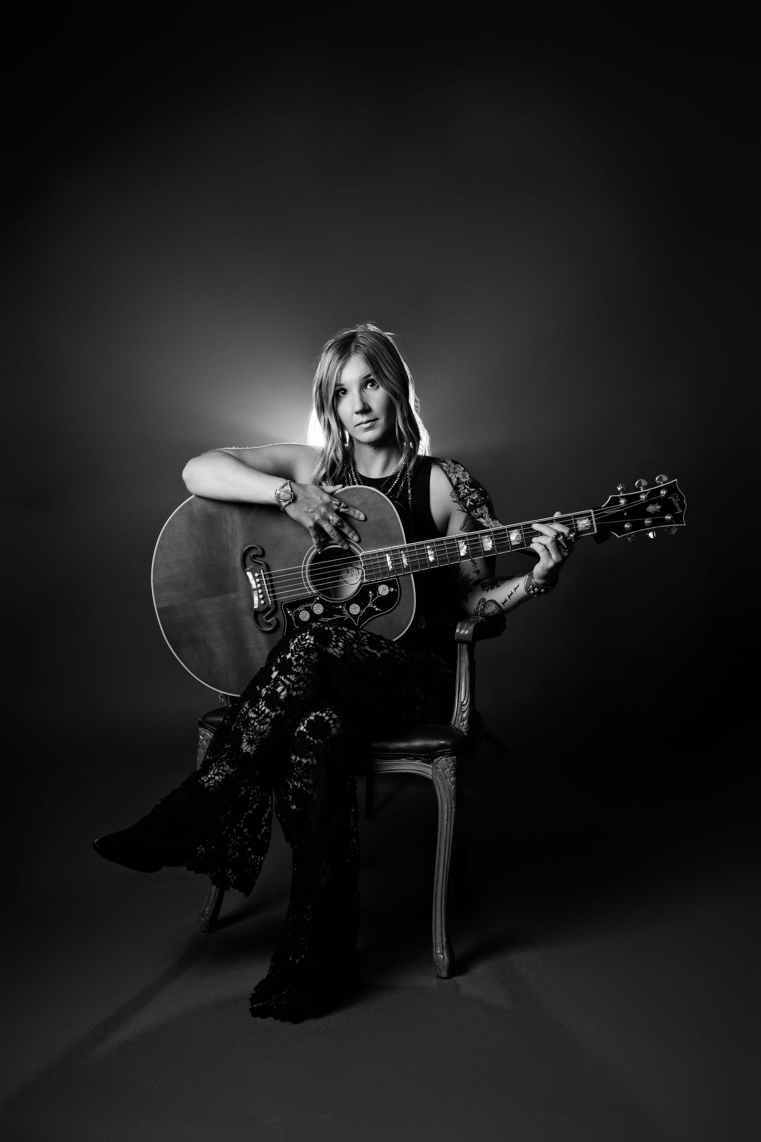 Blonde woman posing with acoustic guitar in chair in elegant black and white studio portrait.