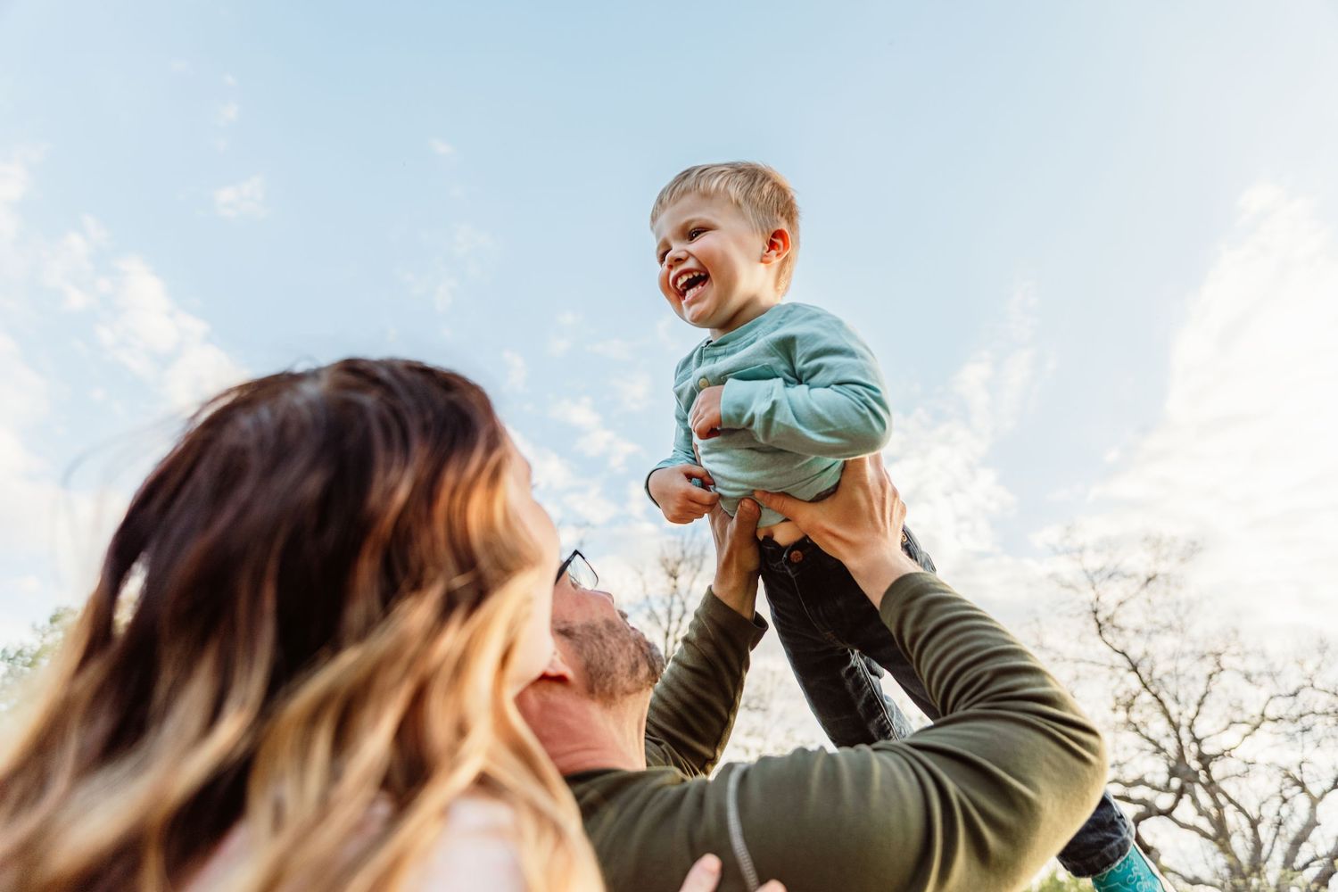 Parents joyfully lifting a laughing toddler in the air outdoors against a blue sky on a sunny day.