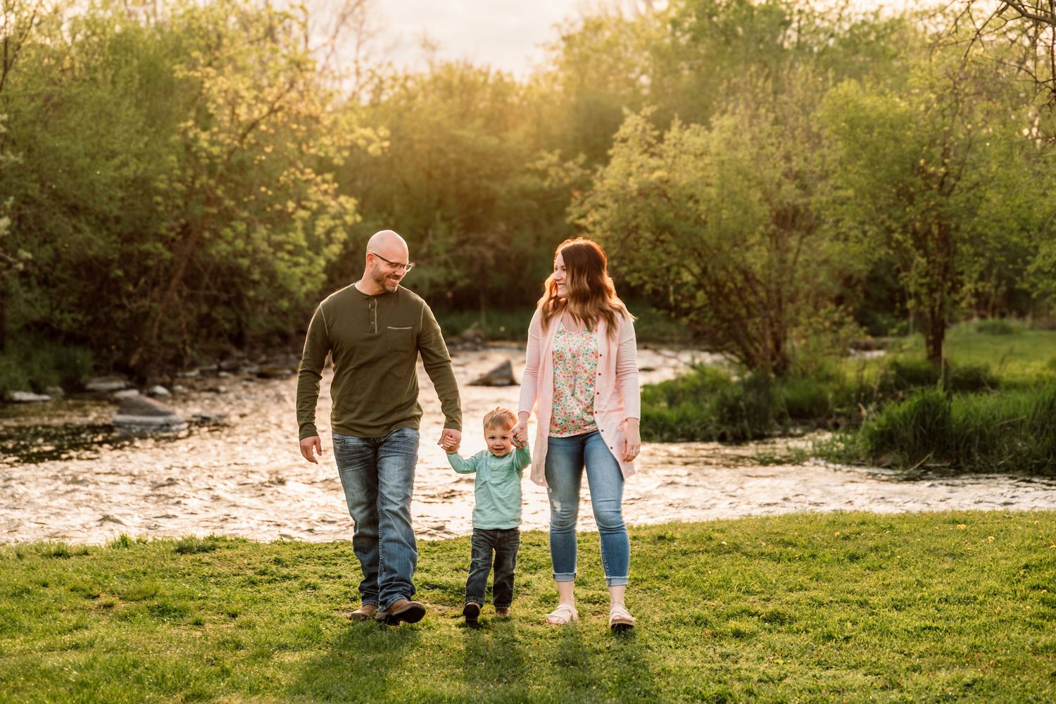Family of three walking hand-in-hand by a sunlit creek surrounded by lush green trees.