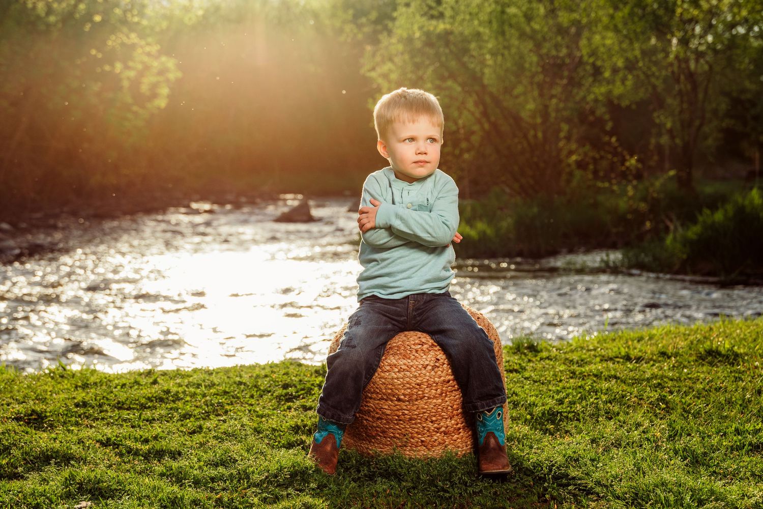 Young boy sitting cross-armed on a wicker basket by a sunlit river, surrounded by lush green grass and trees.