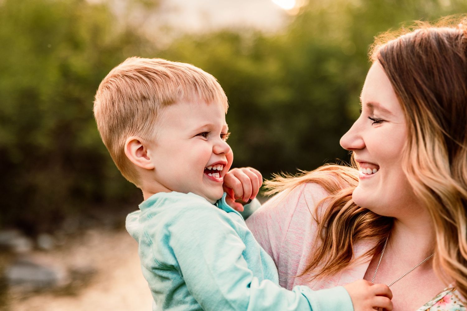 A joyful toddler laughing while being held by a smiling young woman outdoors in warm golden light.