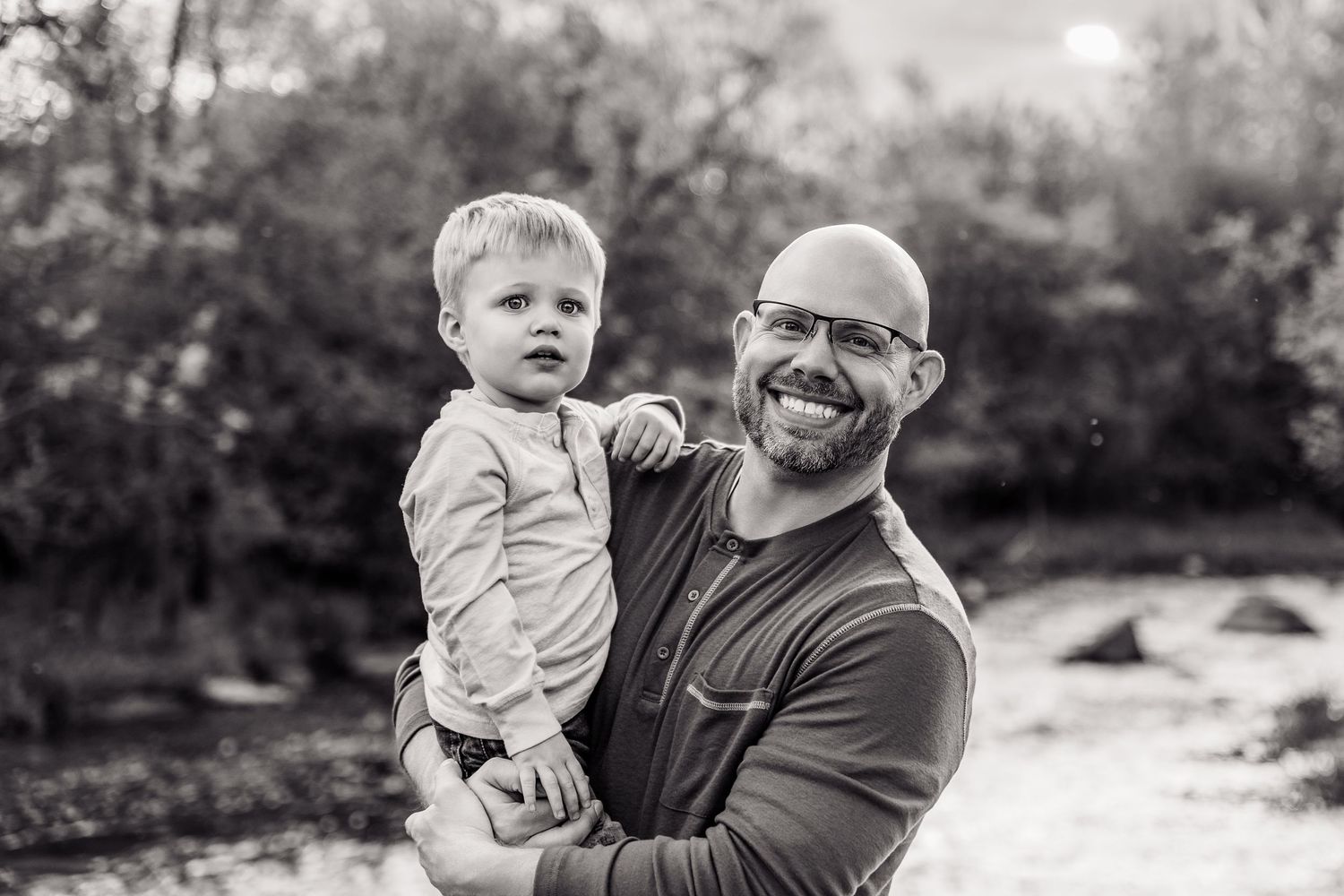 Black and white photo of a smiling bald man with glasses holding a young toddler outdoors near trees.