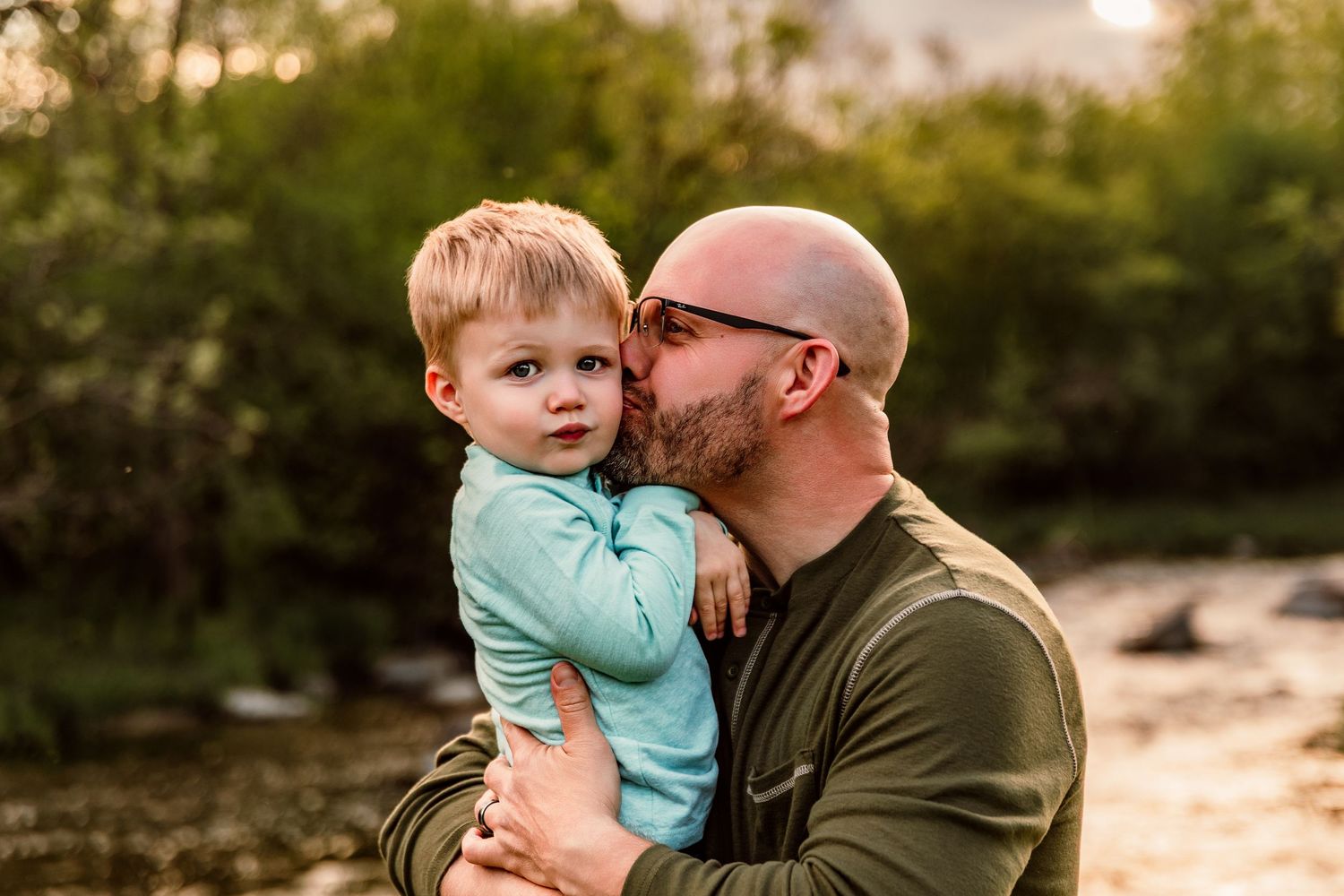 A bald father kisses his young toddler son on the cheek outdoors near a river at golden hour.