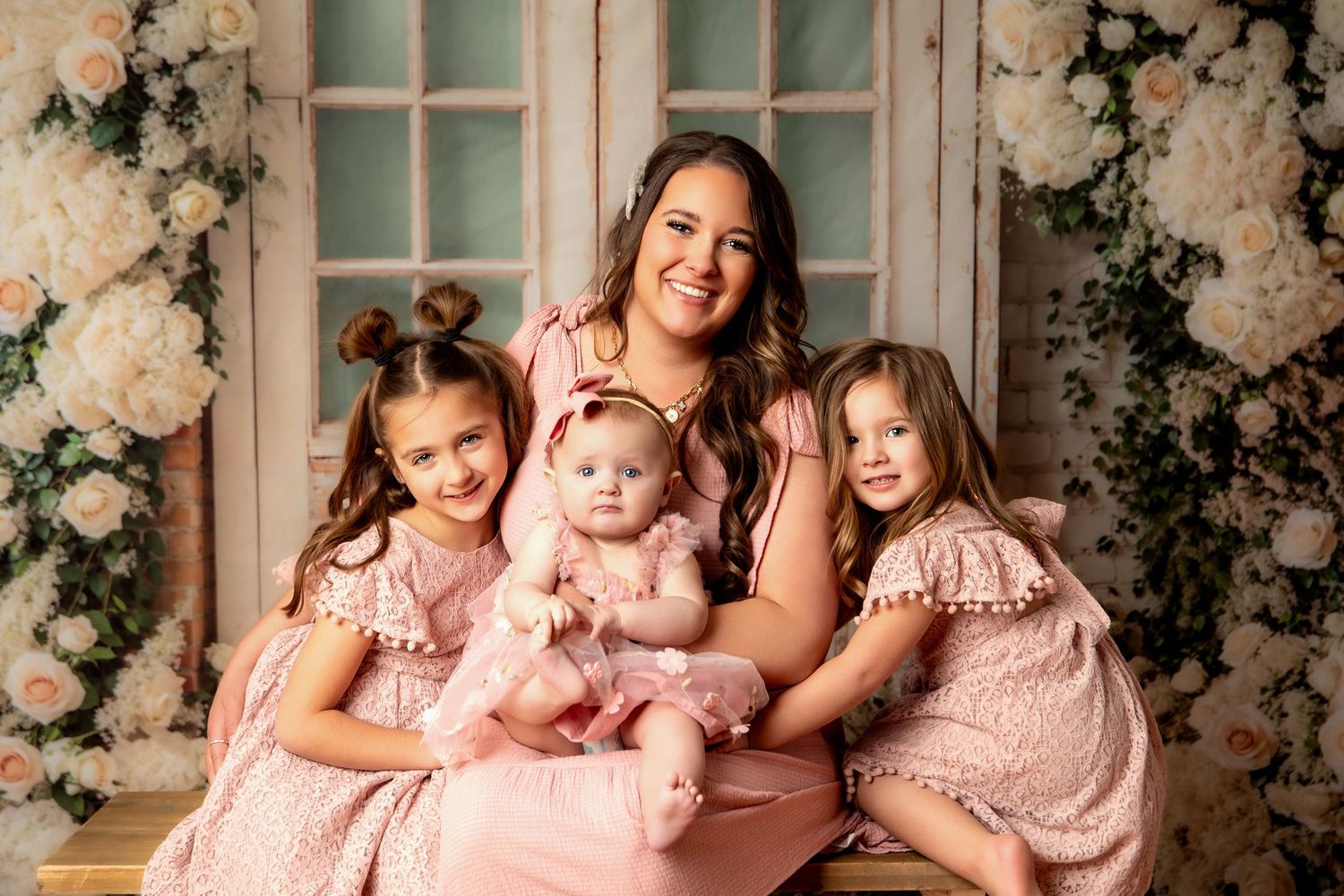 A mother in a pink dress poses with her three young daughters, all wearing matching pink outfits, in a floral studio setting.