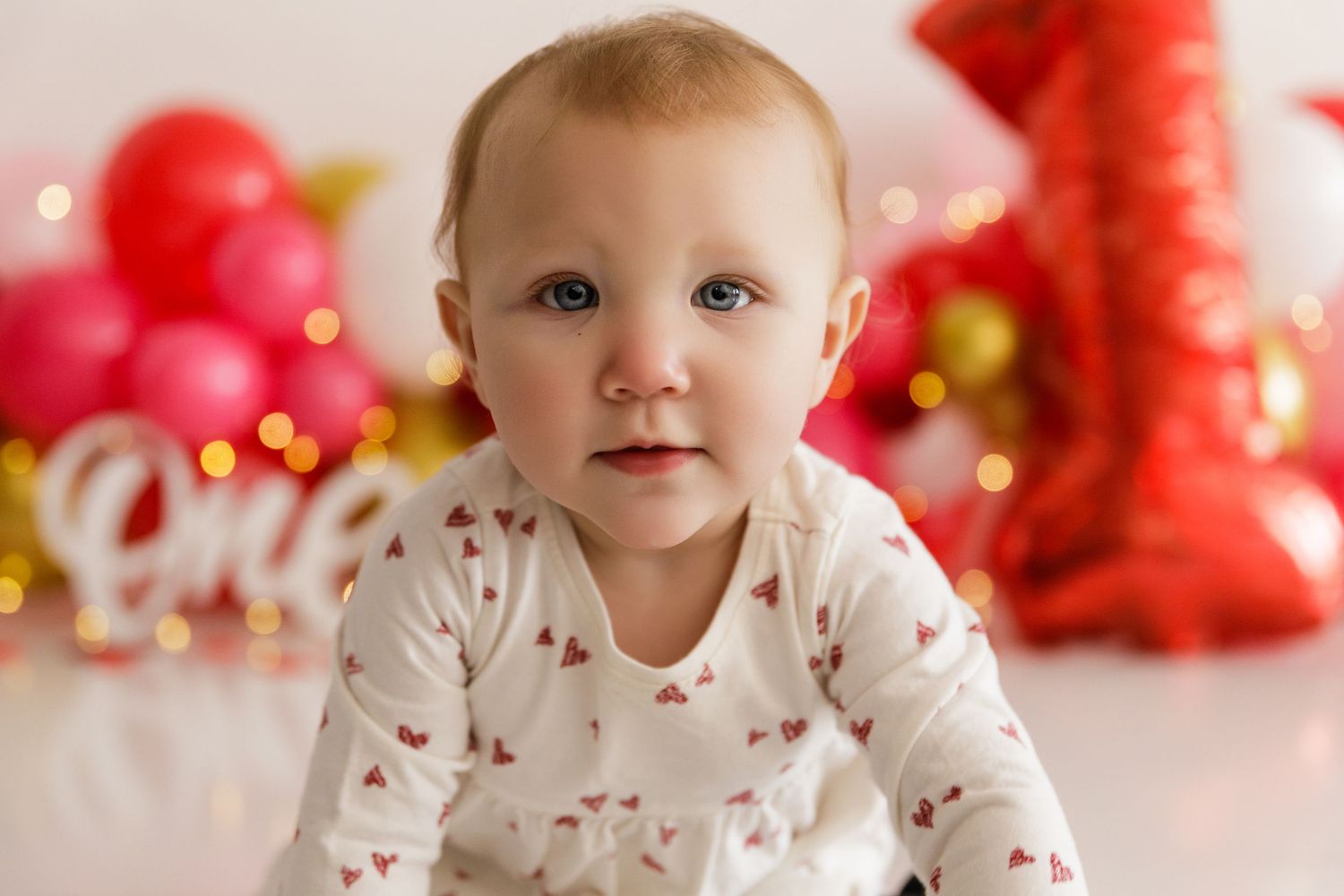 Baby in white patterned outfit sitting against backdrop of red and gold party decorations.