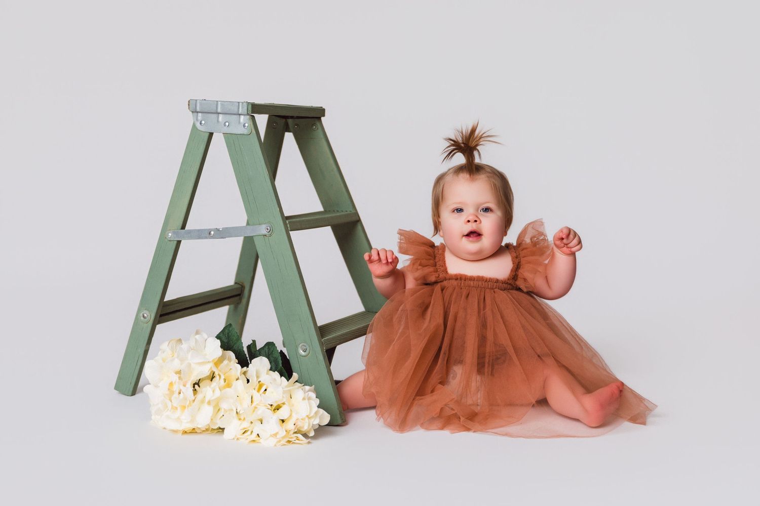 Baby in brown dress reaching towards green ladder with popcorn scattered below.