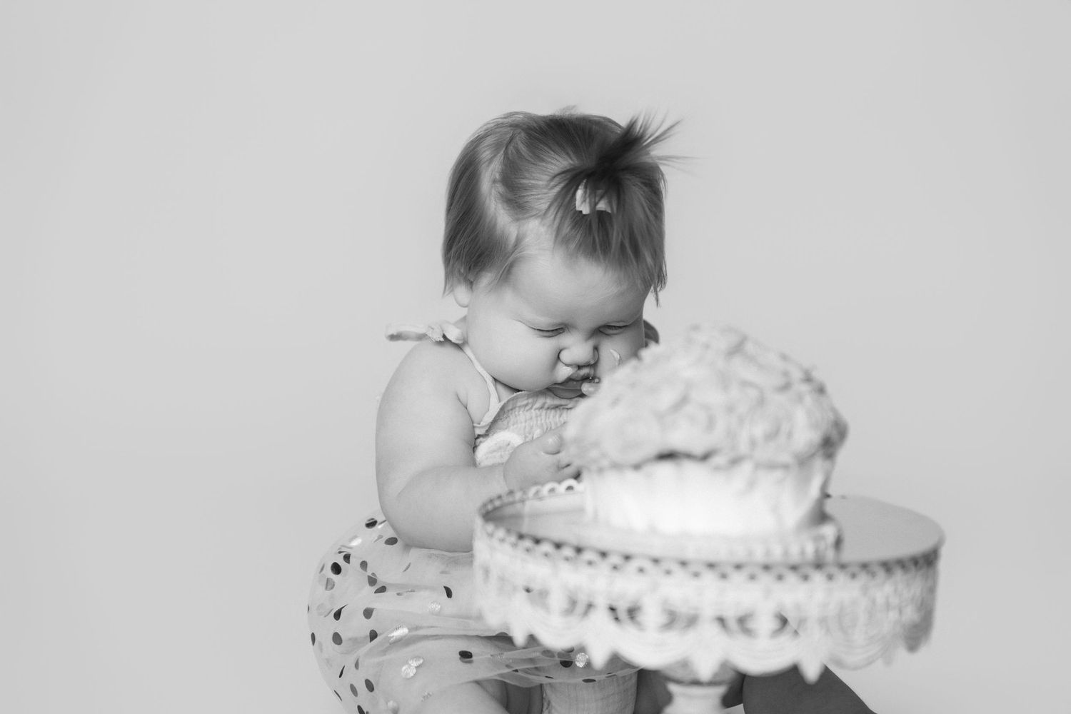 Black and white portrait of baby leaning over to examine cake on ornate pedestal stand.