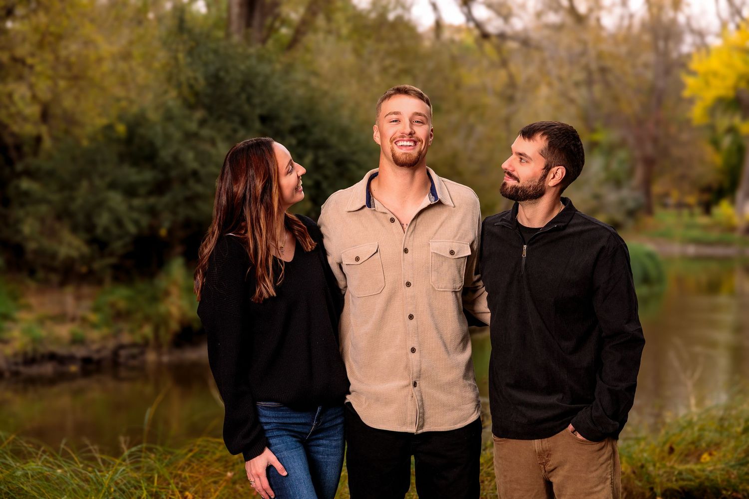 Three people laughing together during a casual outdoor autumn photoshoot near a lake.