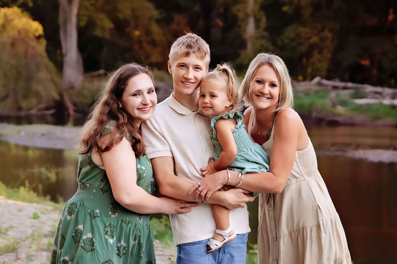 Family wearing coordinated green and white outfits poses together outdoors near a lake during sunset.