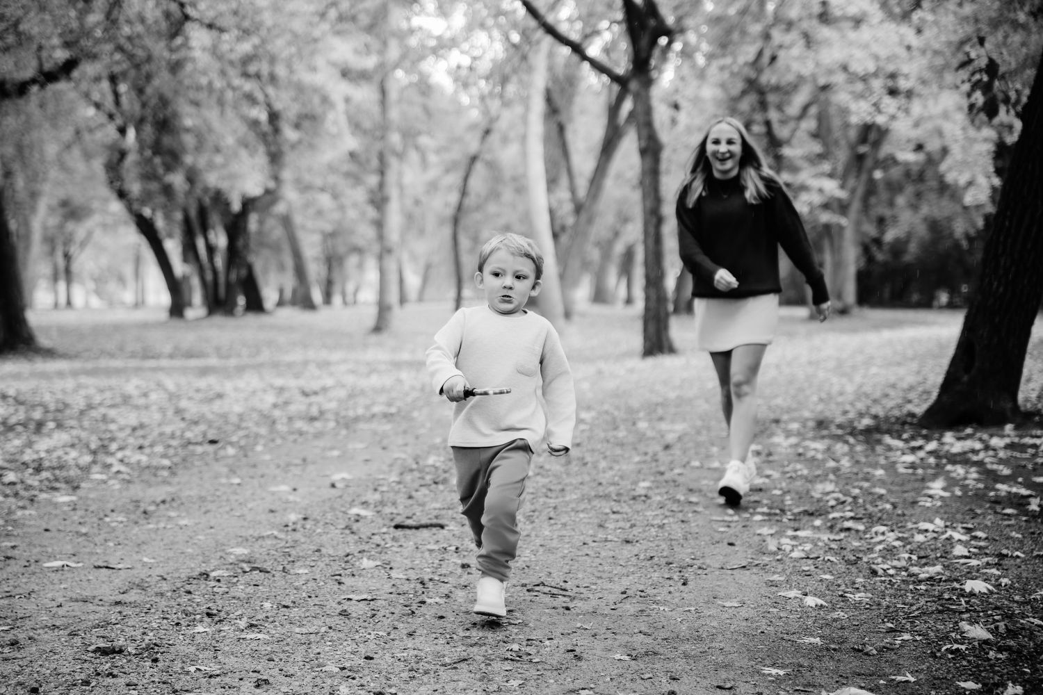 Black and white image showing people walking through a leaf-covered path in a wooded park.