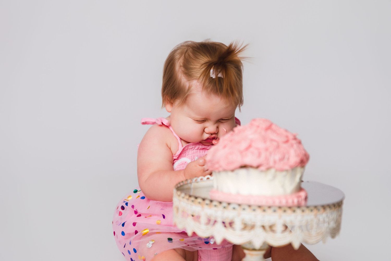 Baby in pink outfit reaches for decorated cake on ornate white stand.