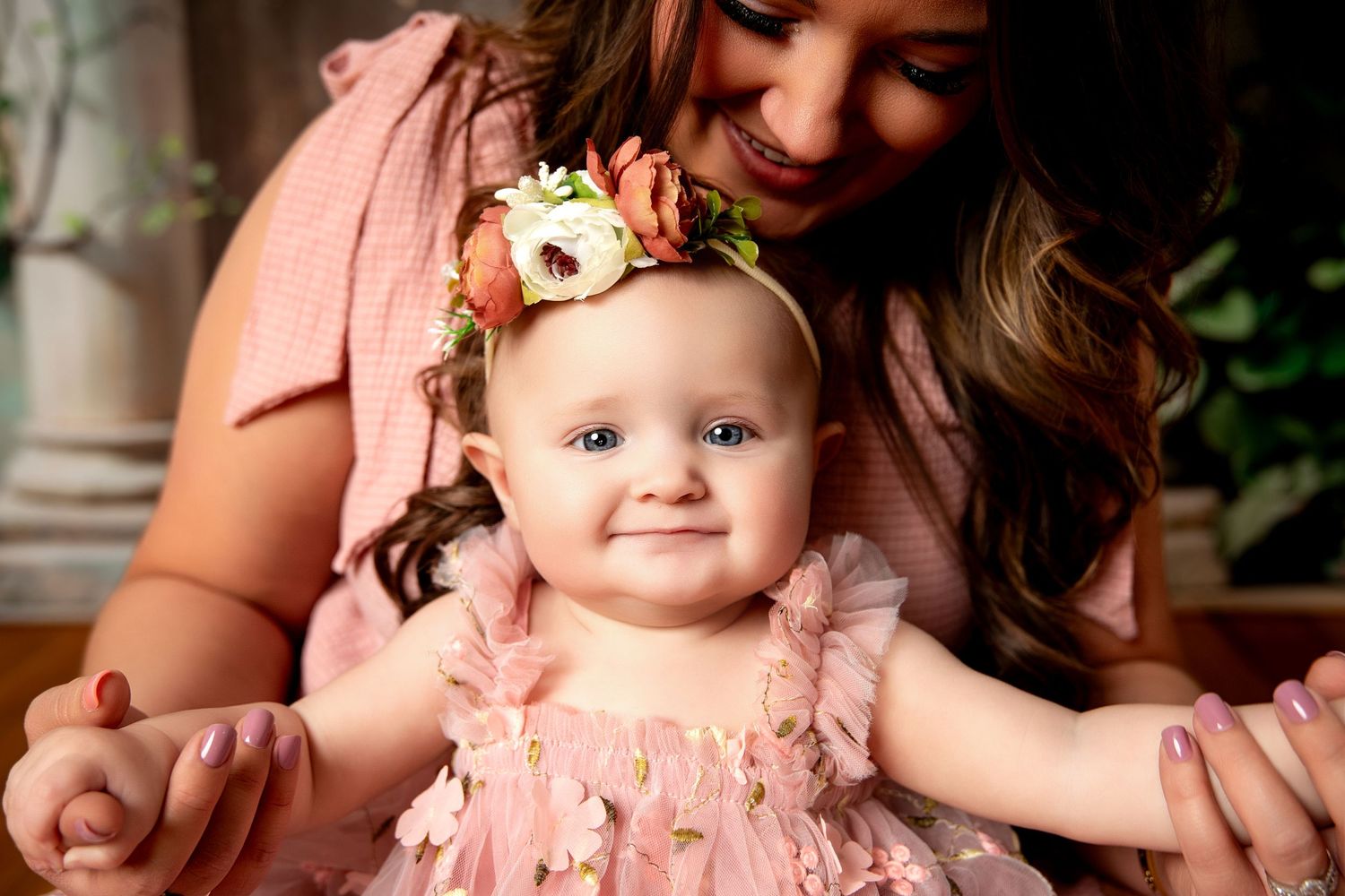 A smiling baby girl in a pink floral dress and flower headband sits with her mother during a professional photo session.