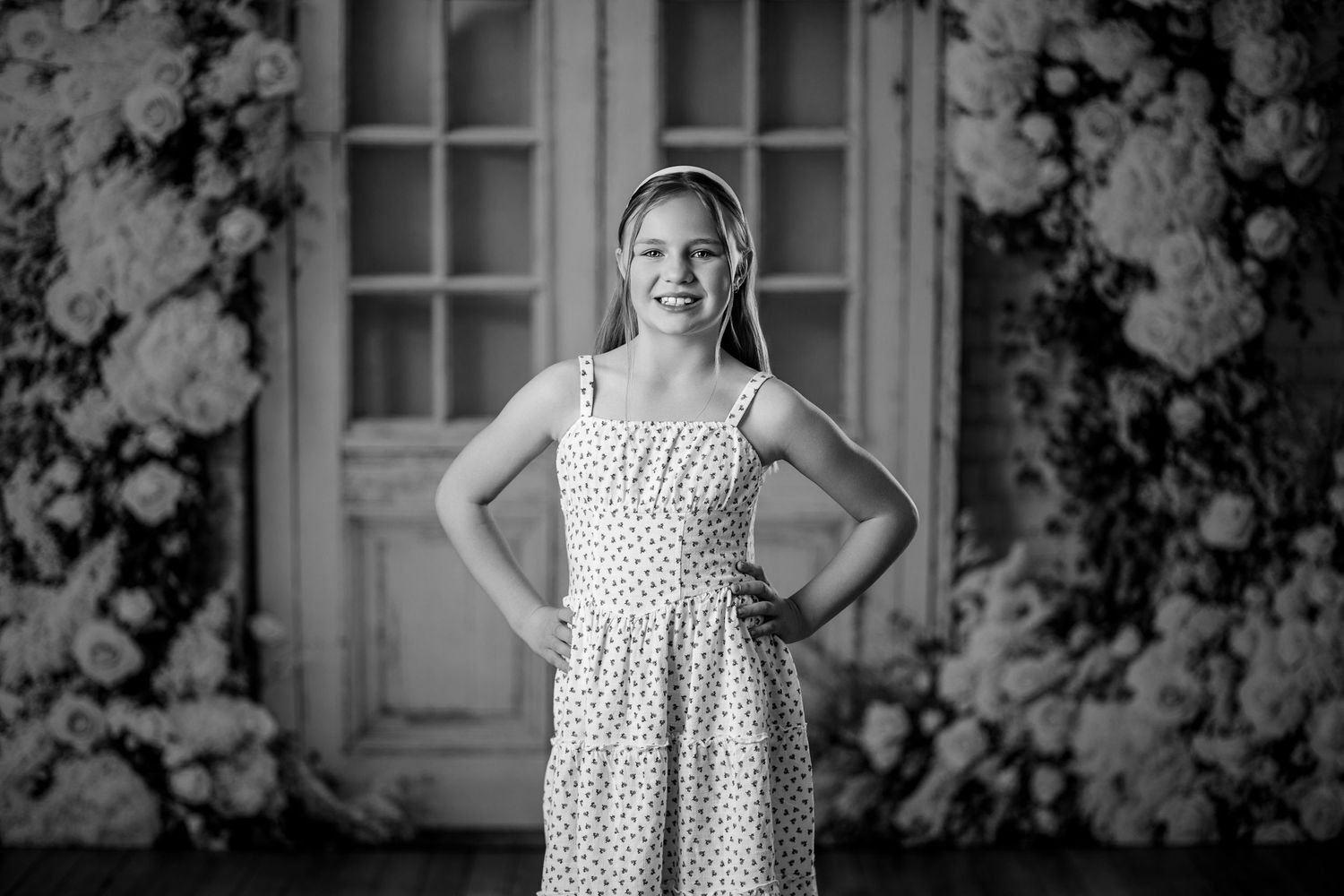 Black and white portrait of a smiling young girl in a polka dot dress standing before a floral and vintage door backdrop.