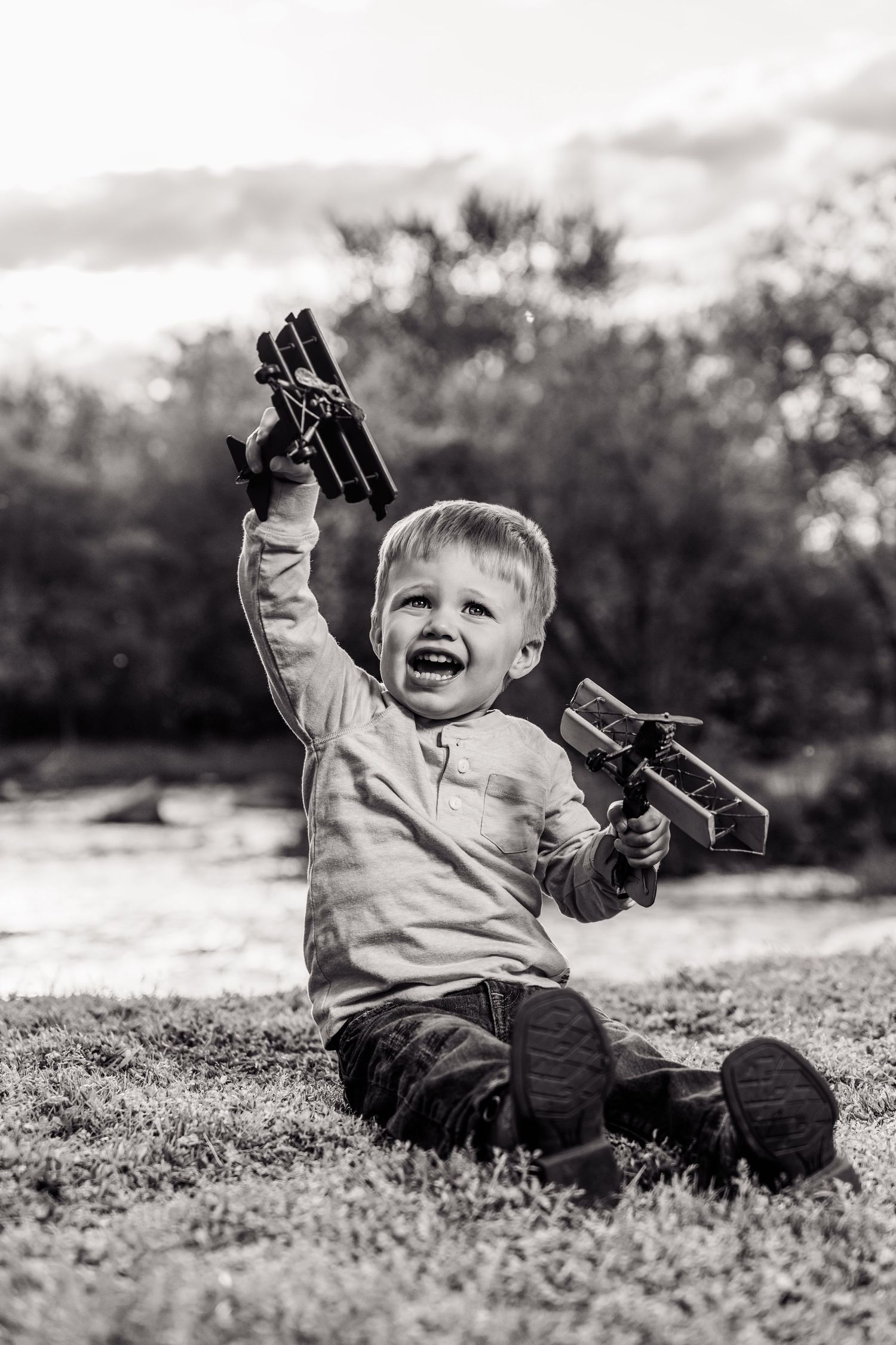 Black and white photo of a joyful toddler boy sitting on grass, gleefully holding up two wooden toy airplanes outdoors.