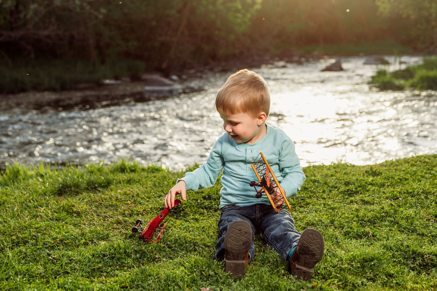 Toddler boy playing with toy planes on green grass beside a sunlit river outdoors.