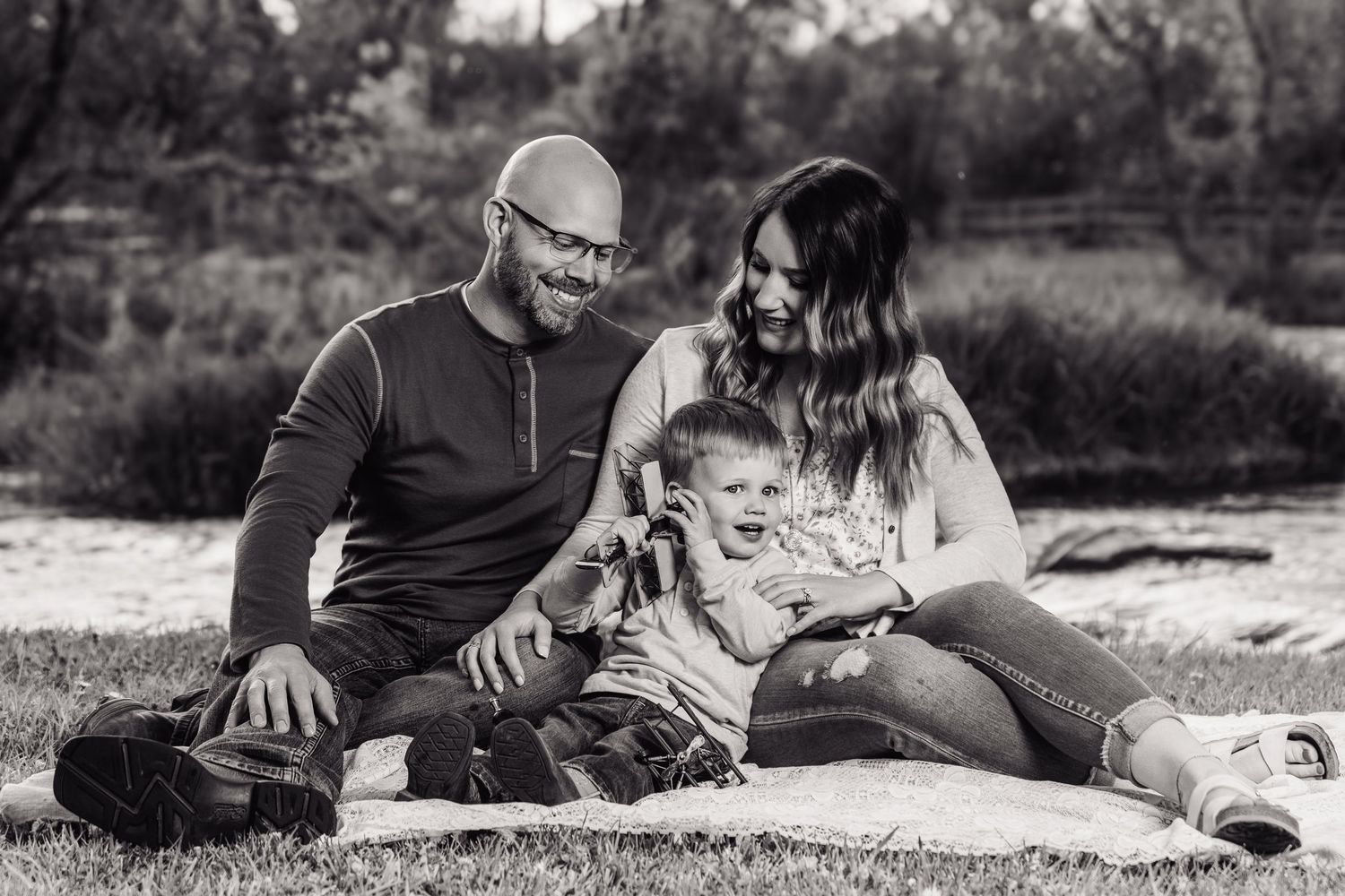 Black and white photo of a happy family sitting on a blanket outdoors, parents smiling with their young child.