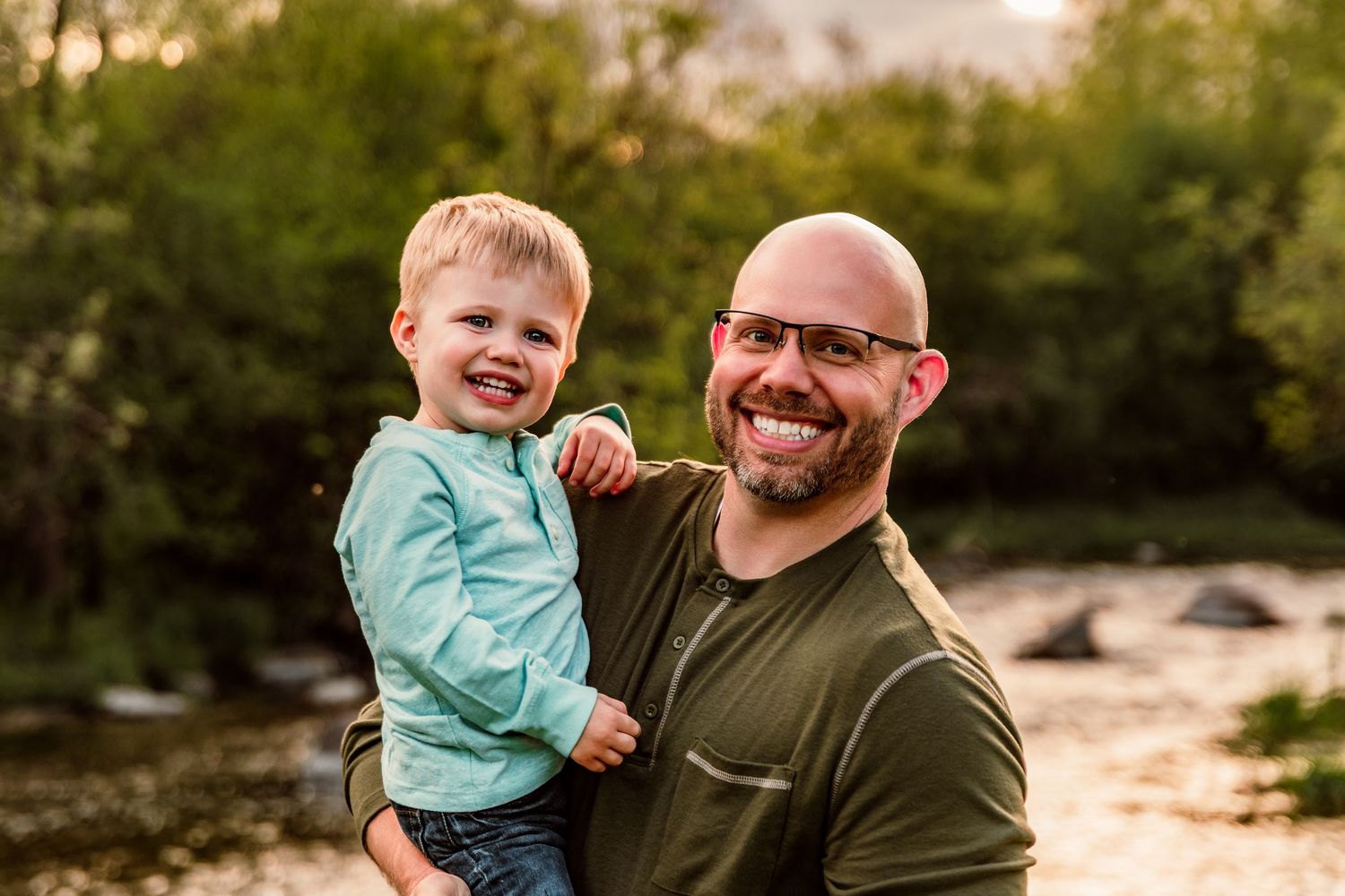 Smiling bald father with glasses holds happy toddler boy outdoors near a river with green trees.