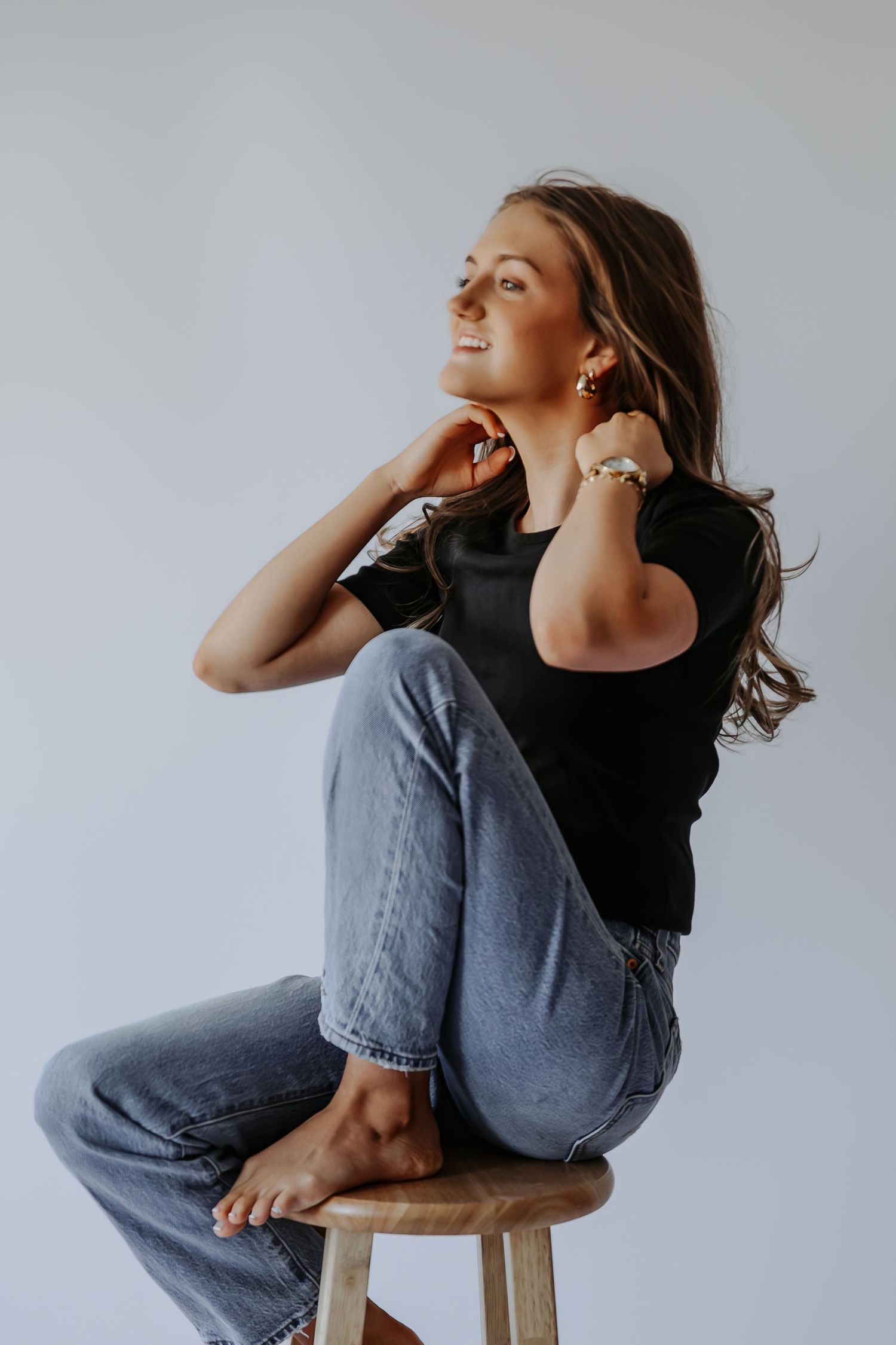Woman in black tee and jeans sitting on stool, smiling with gold earrings and bracelet.