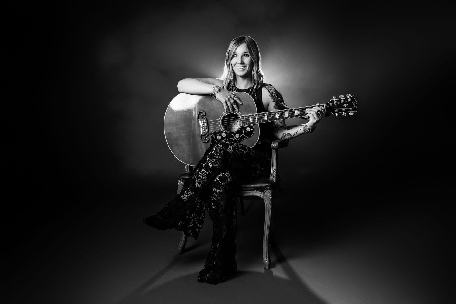 Woman smiling while playing acoustic guitar seated on chair in black and white professional photo.