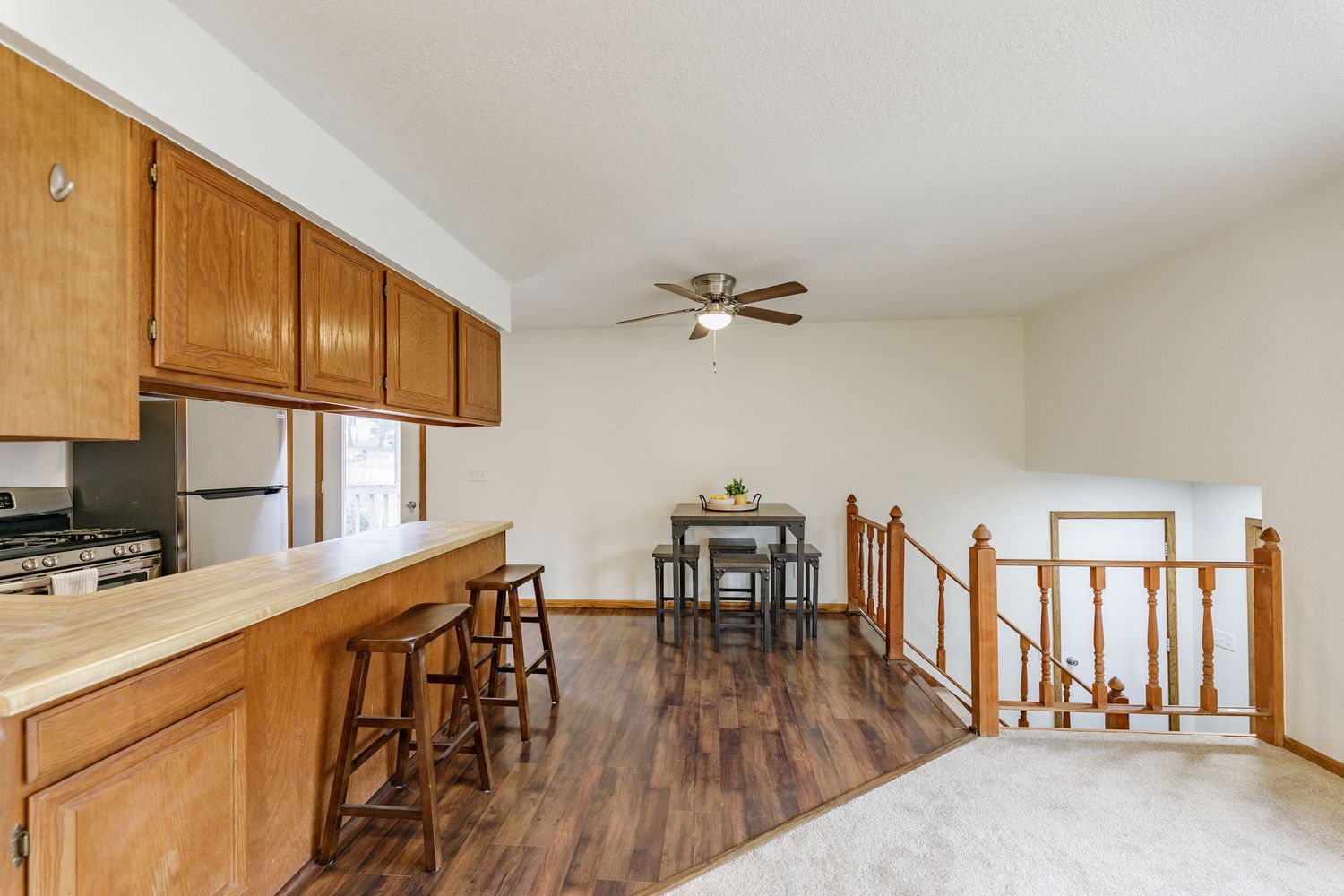 Open-concept kitchen with oak cabinets, bar stools, hardwood floors, ceiling fan, and dining area near staircase railing.